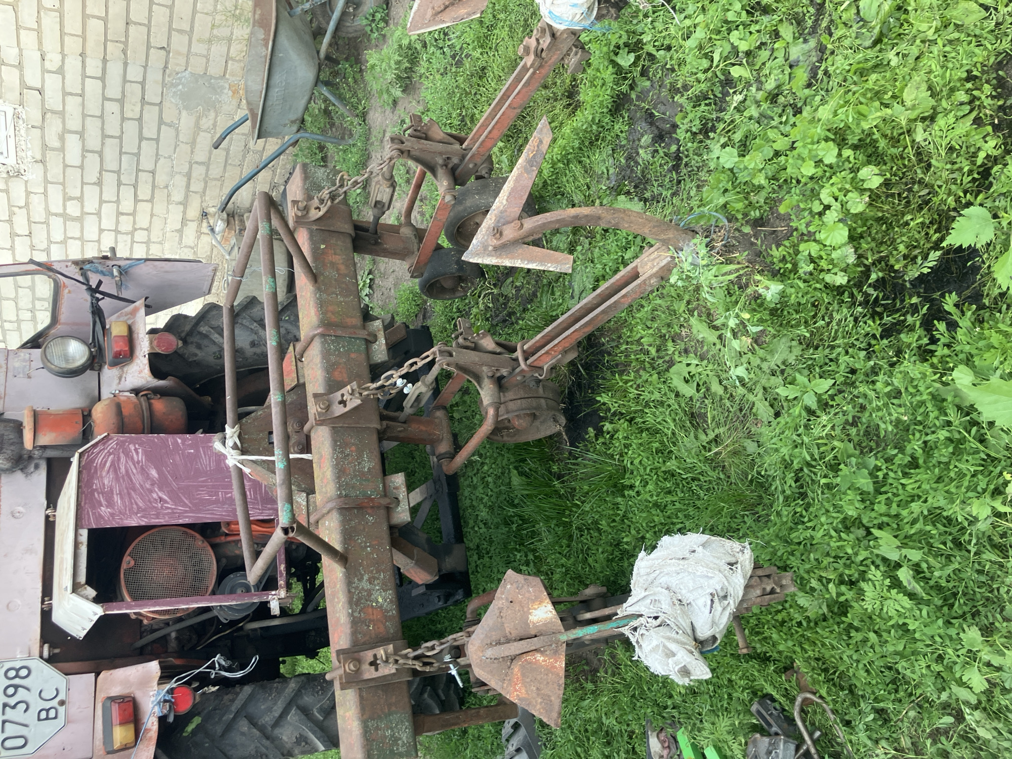An outdoor, medium shot shows agricultural equipment, specifically a tractor and a plow, parked on grassy terrain in Vovkivka, Ukraine. The tractor, partially visible on the left, is faded pink and shows signs of wear. A license plate with the Ukrainian characters "07398 BC" is affixed to its rear. The plow, positioned in the foreground and center, is a rusty, red-brown metal implement with several sharp, metal blades. It is attached to the tractor by thick metal chains. A worn, white sack is tied to one of the plow's components. In the background, a portion of a light-colored brick building is visible on the right, and a weathered grey wheelbarrow stands near it. The ground is covered in dense, green grass with patches of mud. The overall scene depicts agricultural machinery at rest in a rural setting.