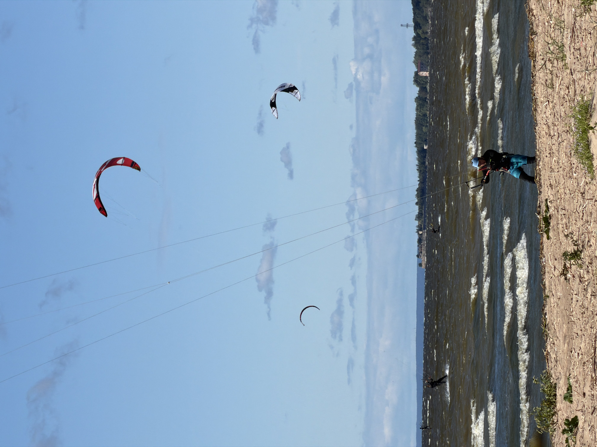 The image depicts a sunny day in Kronstadt, Russia, with a person kitesurfing on a choppy body of water. The sky is a clear blue with a few scattered clouds. Several kites are visible in the sky, one red and white, and another black and white, indicating multiple kitesurfers are present or have been. The kites are connected by lines to the kitesurfer(s), who are actively engaged in the sport.
The foreground shows a sandy shore with some sparse vegetation. The water is rough with visible waves, suggesting windy conditions suitable for kitesurfing. In the background, a tree line is visible along the horizon, with some structures, possibly buildings, peeking through the trees. The sun appears to be high in the sky, suggesting it is midday.
One kitesurfer is clearly visible on the water, wearing a black wetsuit and blue shorts, and a helmet. They are in motion, with spray coming off the water around them. Other figures are visible in the distance on the water, also appearing to be kitesurfing. The overall scene suggests an active and recreational day at the coast. There is no visible text in the image.