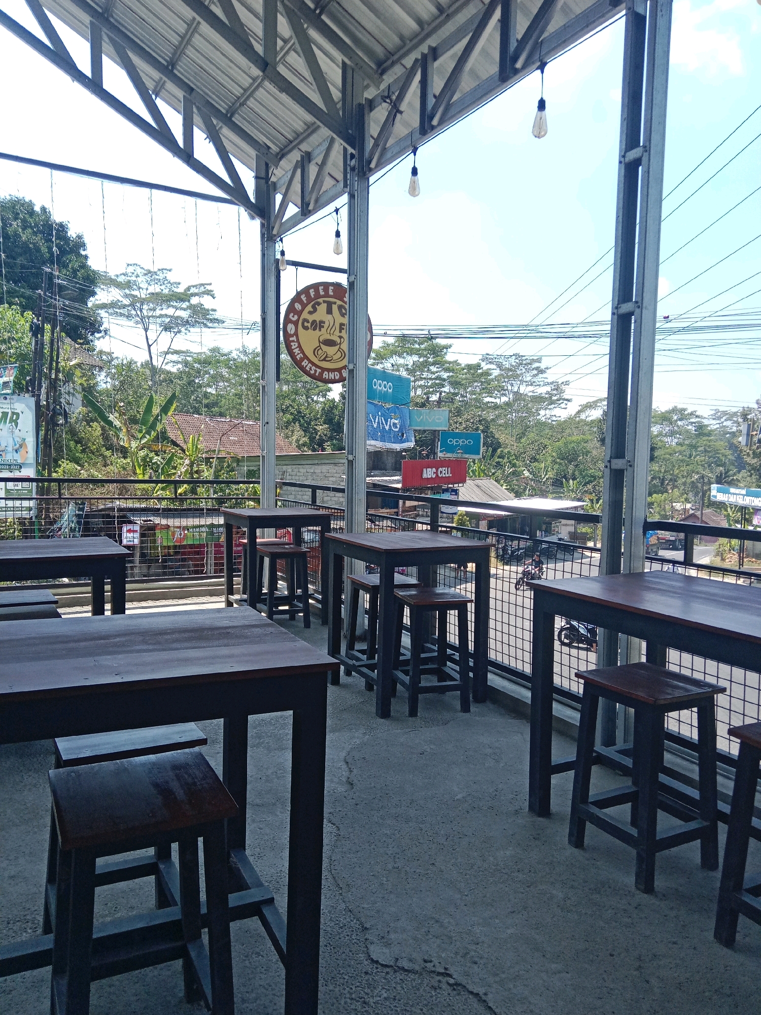 The image depicts an outdoor seating area of a coffee shop, likely located in Tanjunganom, Indonesia, as suggested by the context. The foreground features several dark wooden tables and stools set on a concrete floor. Beyond the seating area, a metal railing separates the patio from the street. In the background, there are various buildings, trees, and utility wires, characteristic of a semi-urban or rural Indonesian setting. Signs for "COFFEE STOP," "OPPO," and "VIVO" are visible, indicating commercial activity. The sky is bright and clear, suggesting daytime. There are no people clearly visible in the immediate seating area, but a person on a motorcycle can be seen in the background on the street. The overall atmosphere is casual and open-air.