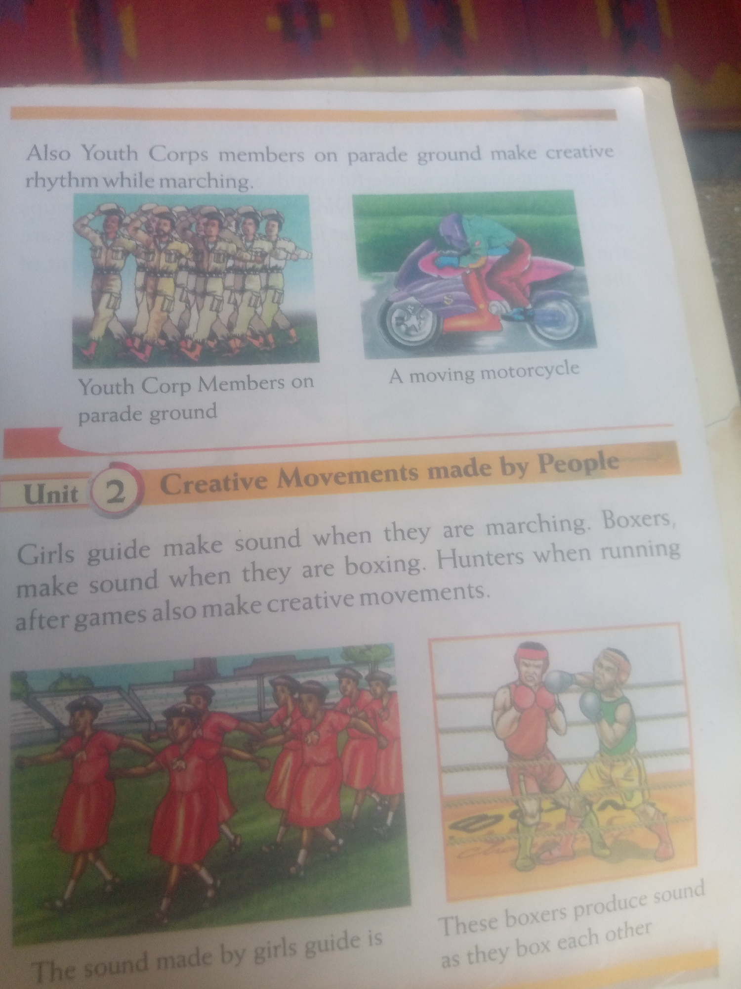 The image displays a page from a book. At the top, a caption reads "Also Youth Corps members on parade ground make creative rhythm while marching." Below this are two illustrations. The left illustration shows a group of people in uniform marching. The right illustration depicts a person riding a motorcycle. Beneath the illustrations, text reads "Youth Corp Members on parade ground" and "A moving motorcycle."
A horizontal orange bar separates the top section from the main content. The bar contains the text "Unit 2 Creative Movements made by People."
The main text states, "Girls guide make sound when they are marching. Boxers, make sound when they are boxing. Hunters when running after games also make creative movements."
Below this text are two more illustrations. The left illustration shows a group of girls in red dresses, possibly a drill or dance routine. The right illustration shows two boxers in a ring, engaged in a match.
At the bottom, text beneath the girls' illustration reads, "The sound made by girls guide is". Text beneath the boxers' illustration reads, "These boxers produce sound as they box each other".
The location in the city of Jauro Yinu, Nigeria, is not mentioned or depicted in this image.