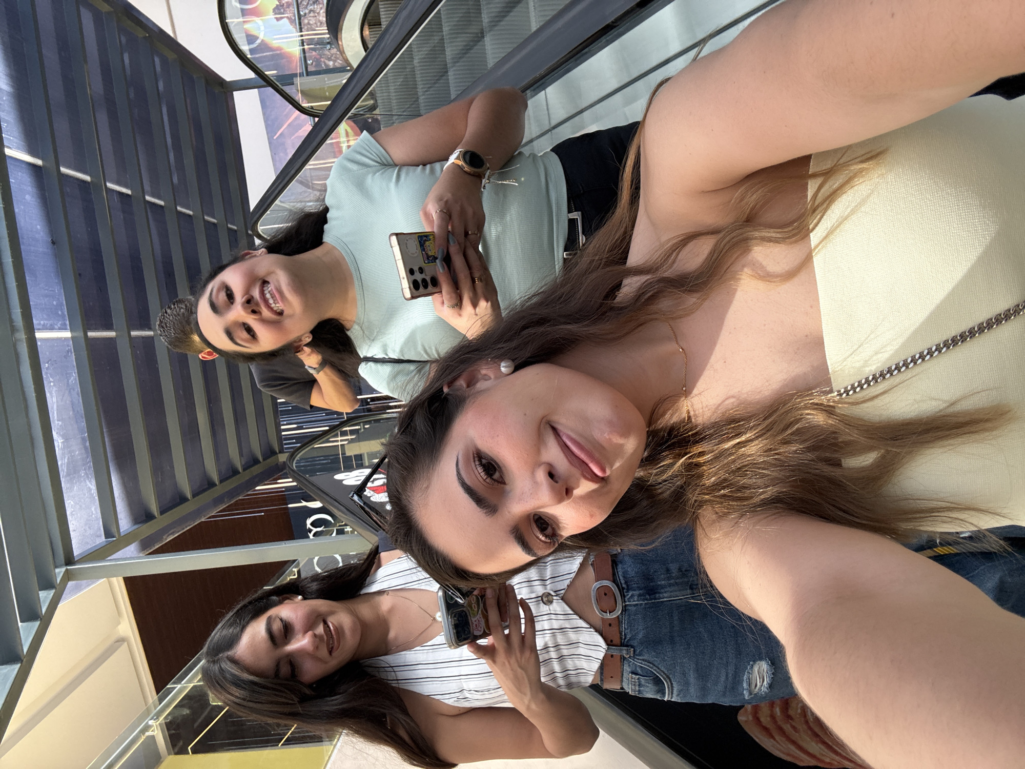 Three women are taking a selfie on an escalator. The location appears to be indoors, possibly a mall, as suggested by the escalator and interior architectural features. The women are holding phones, suggesting they are taking the photo themselves. One woman is sticking her tongue out. They are located in San Bernardino Tlaxcalancingo, Mexico.