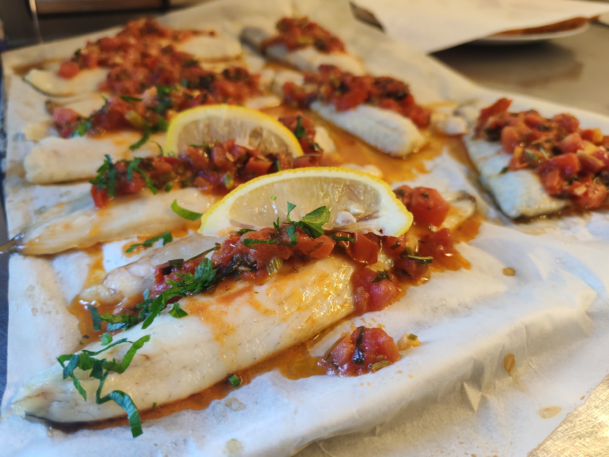 The image shows several pieces of white fish, each topped with a diced tomato and herb mixture, arranged on a sheet of parchment paper. Slices of lemon are placed on some of the fish fillets. The parchment paper is on a metallic surface. The scene appears to be a close-up shot of prepared food, likely ready to be cooked or served. The overall impression is one of fresh, homemade cuisine. There is no indication of the city of La Spezia, Italy.