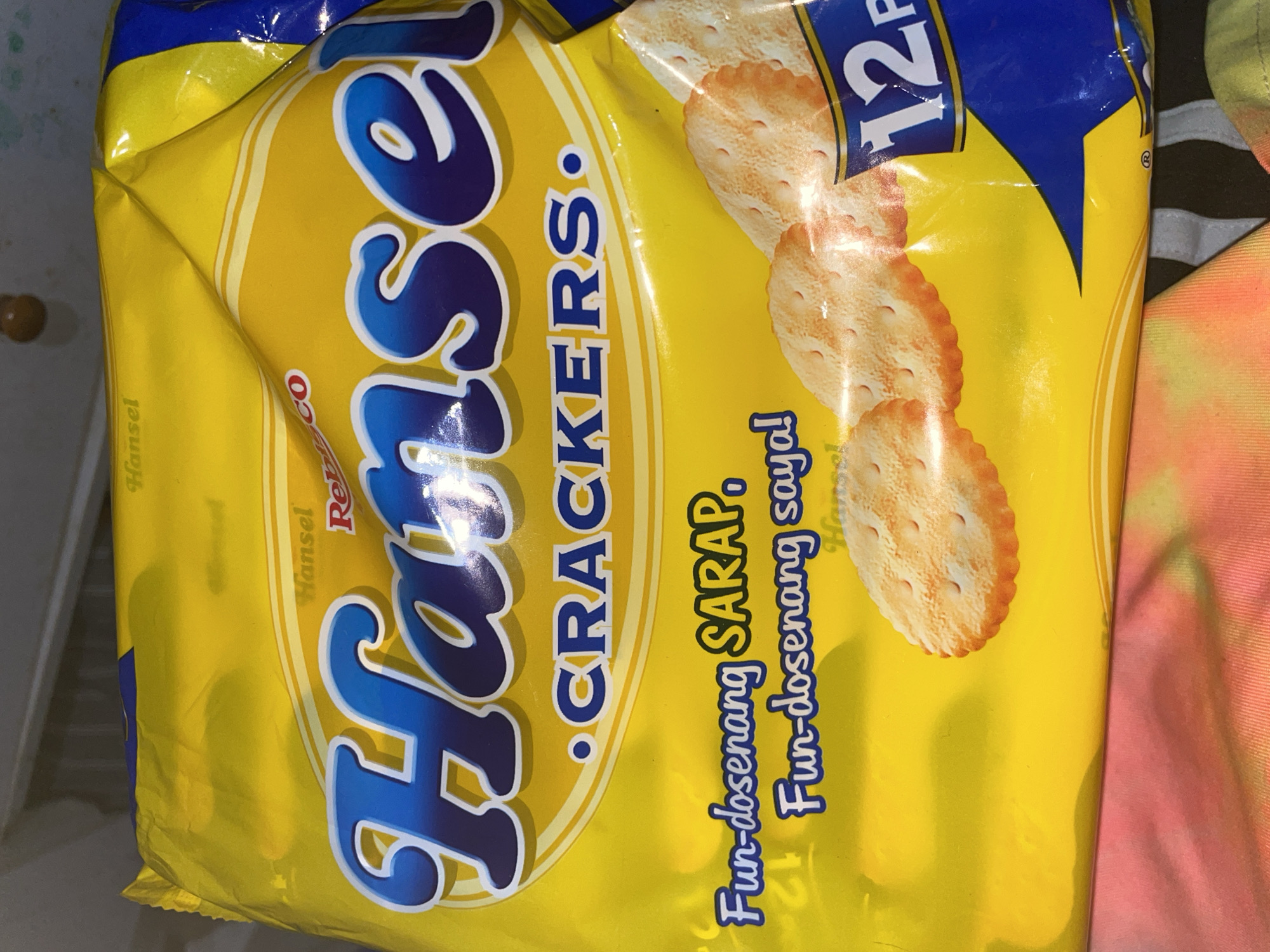 A close-up, eye-level shot shows a brightly lit yellow package of Hansel Crackers, a product of Universal Robina Corporation. The packaging prominently displays the brand name "Hansel" in large, blue, stylized lettering with white and purple outlines. Below the brand name, "CRACKERS" is printed in a similar font. Text in a slightly smaller, blue font reads "Fun-dosenang SARAP" and "Fun-dosenang saya!", which translates to "Fun-dosenang DELICIOUS" and "Fun-dosenang fun!". The packaging is slightly creased, indicating it's likely a product in a store or a home. In the lower right corner, the number "12" is visible within a blue banner, likely indicating a quantity or pack size. Scattered across the yellow background are several round, golden-brown crackers with visible perforations. The bottom of the image shows a blurred colorful textile, possibly clothing or a tablecloth. No specific location in Davao City is identifiable from the image.