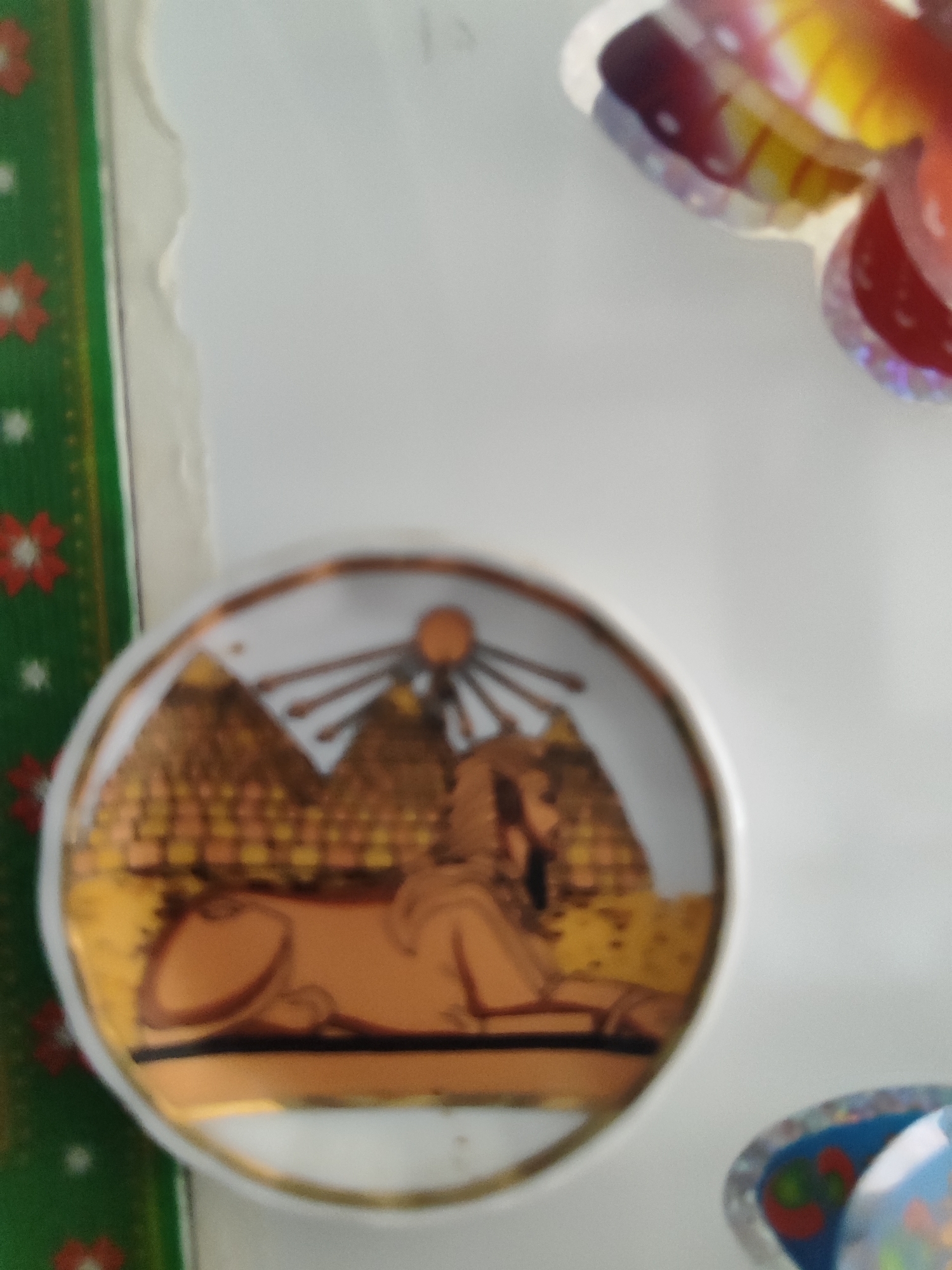 The image shows a close-up of a round, decorative plate featuring an illustration of the Great Sphinx and pyramids of Giza, Egypt. The plate has a gold rim and the illustration is in gold and orange tones. Behind the plate, on the left side of the image, is a green border with red and white floral patterns. To the right of the plate, a blurry, colorful object, possibly another decoration or a piece of candy, is visible. The background is a plain white surface.