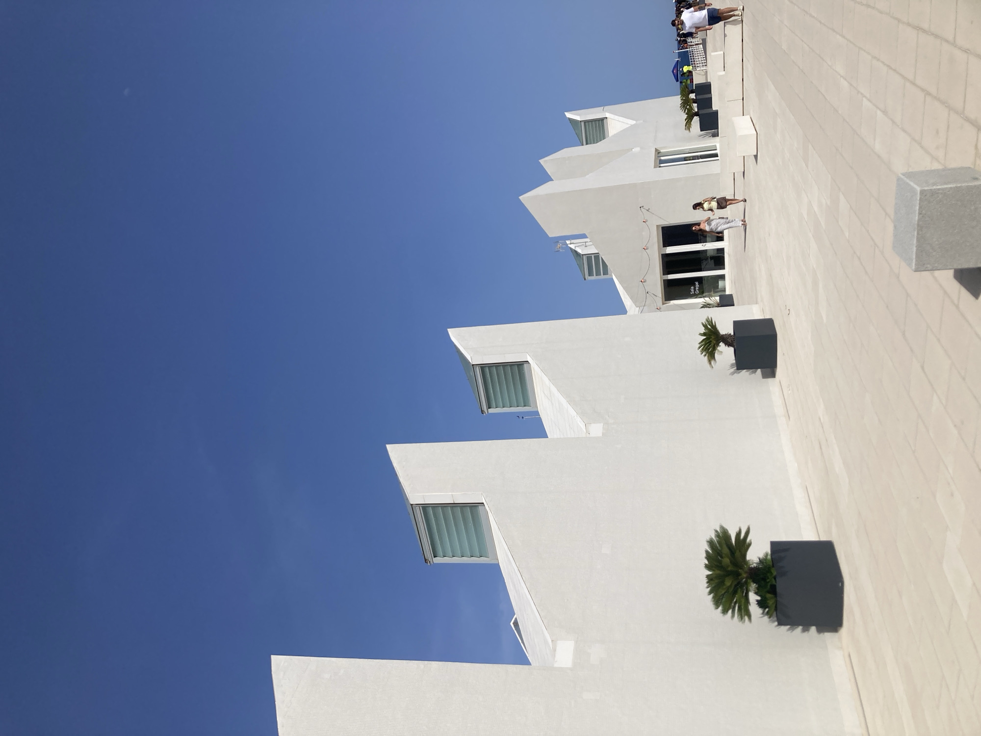 This is a wide-angle, eye-level shot of a modern, white, geometric building with sharp angles and a flat roof against a clear blue sky. The building has several louvered windows positioned at an angle on its upper sections. In front of the building is a paved area with light-colored square tiles. Several dark gray planters with small palm trees are strategically placed along the building's facade. Two women in casual attire are walking towards the building entrance, which features glass doors with text that appears to read "Sala Griegal." Further in the distance, a few other people are visible walking on the tiled area, and a yellow barrier is present. The architectural style and the seaside setting, with a hint of beach activity in the far right background, suggest this building could be located in a coastal area of Barcelona, Spain, possibly serving as a cultural or recreational facility.