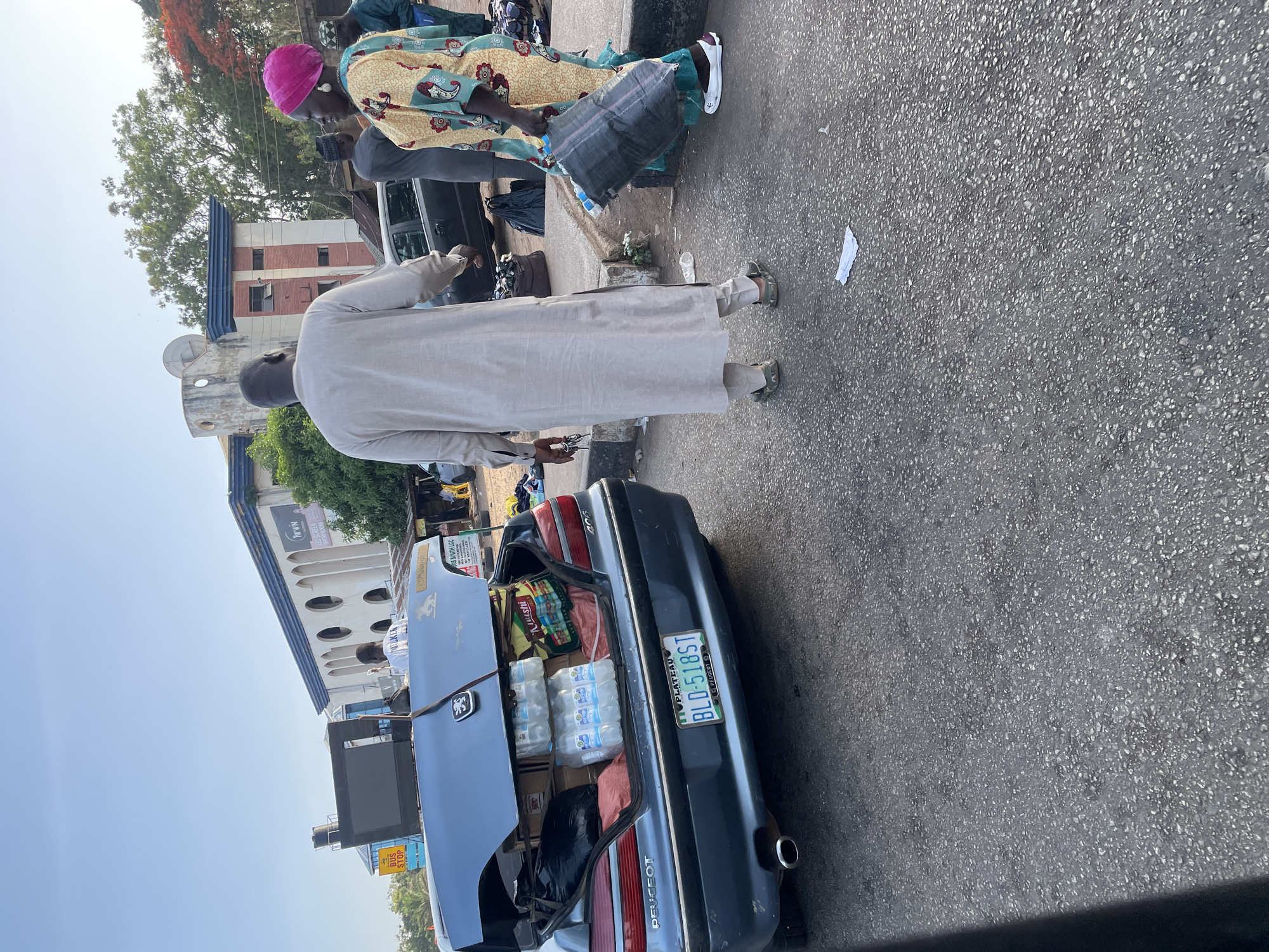 The image captures a street scene in Jos, Nigeria, featuring a dark blue Peugeot 406 sedan with its trunk open. The trunk is packed with goods, including bottled water and bags. A man in a long, light-colored traditional robe stands by the car. Several people are nearby, one wearing a pink head covering and a patterned dress. The license plate is visible and reads "BLD-518 ST" and "Plateau." A building with a clock tower is in the background. The setting appears to be a roadside with pedestrian activity.