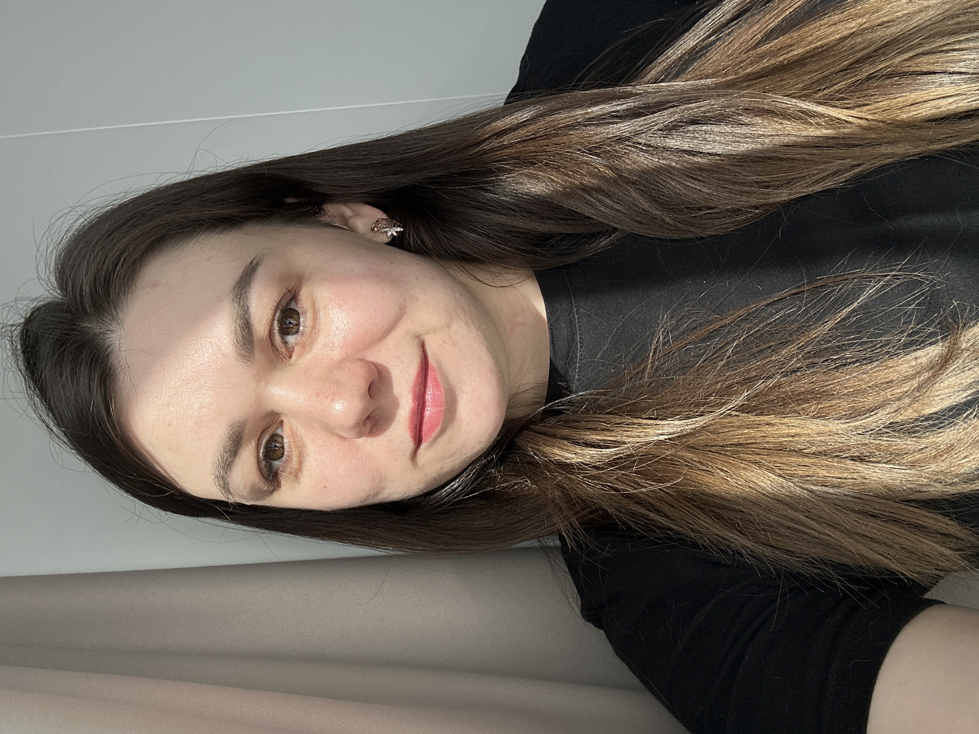 A medium shot depicts an adult female indoors in the city of Chaikovsky, Russia. The subject has a light complexion, brown eyes, and long, straight to wavy hair that is dark brown at the roots and transitions to a lighter brown or auburn shade towards the ends. She is wearing a black, presumably crew-neck, top. A gold-toned earring with an intricate, possibly floral, design is visible in her right ear. Her lips are tinted with a light pink cosmetic. The subject is looking directly at the camera with a subtle, closed-mouth smile. Strong natural light illuminates the left side of her face and hair, creating a distinct shadow line across her features. The immediate background on the left consists of a light-colored, vertically pleated curtain or drape, while the right side reveals a plain light grey wall.