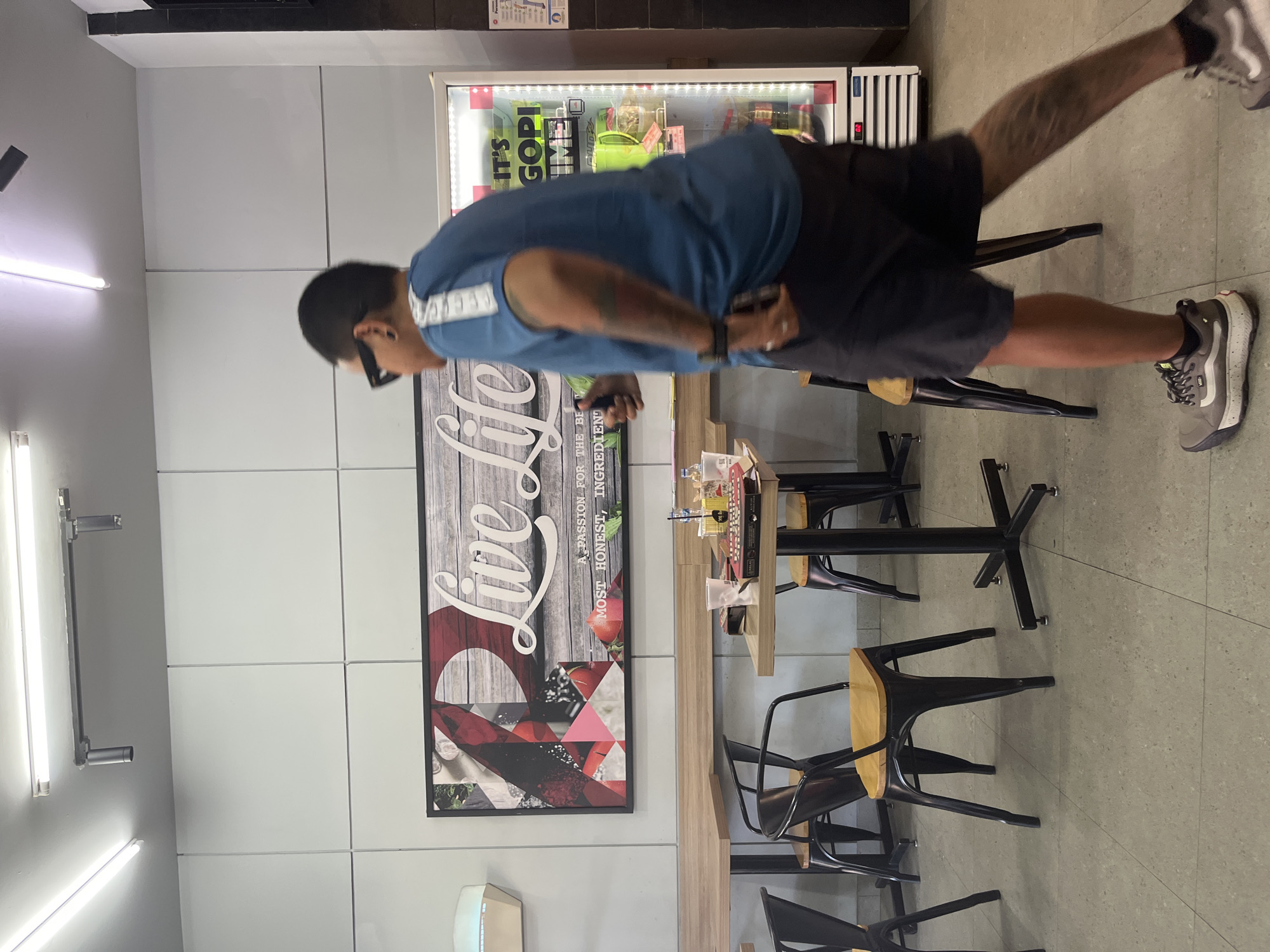 In the city of Kuta Selatan, Indonesia, an individual wearing a blue t-shirt and dark shorts is captured mid-stride within an indoor establishment, possibly a cafe or restaurant. The person, seen from the side, has dark hair, sunglasses, and tattoos visible on their arm. They are walking past a glass-fronted refrigerator displaying various beverages. In the background, a large framed artwork with the text "Live Life" is mounted on a light-colored tiled wall. Several tables and chairs, primarily black with wooden seats, are arranged throughout the space, indicating a seating area for customers. The overall environment appears to be a well-lit commercial interior with a tiled floor.