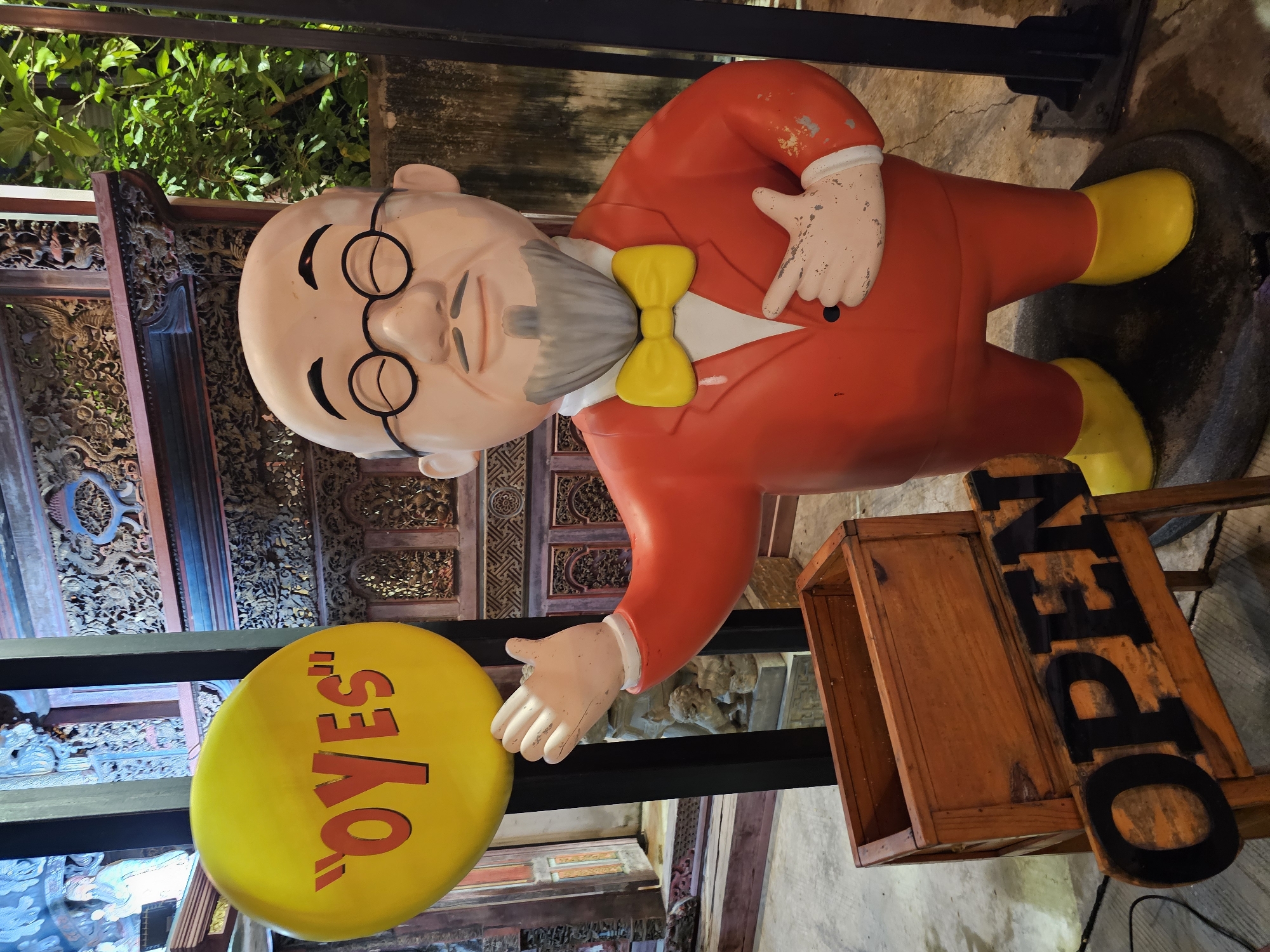 A statue of a bald man wearing a red suit, yellow bowtie, and glasses stands to the right of a yellow circular sign with "OYES" written in red. In front of the statue is a wooden box and a sign with the word "OPEN" in black letters. The background appears to be an outdoor market in Bangkok, Thailand, with intricate wooden carvings and greenery.