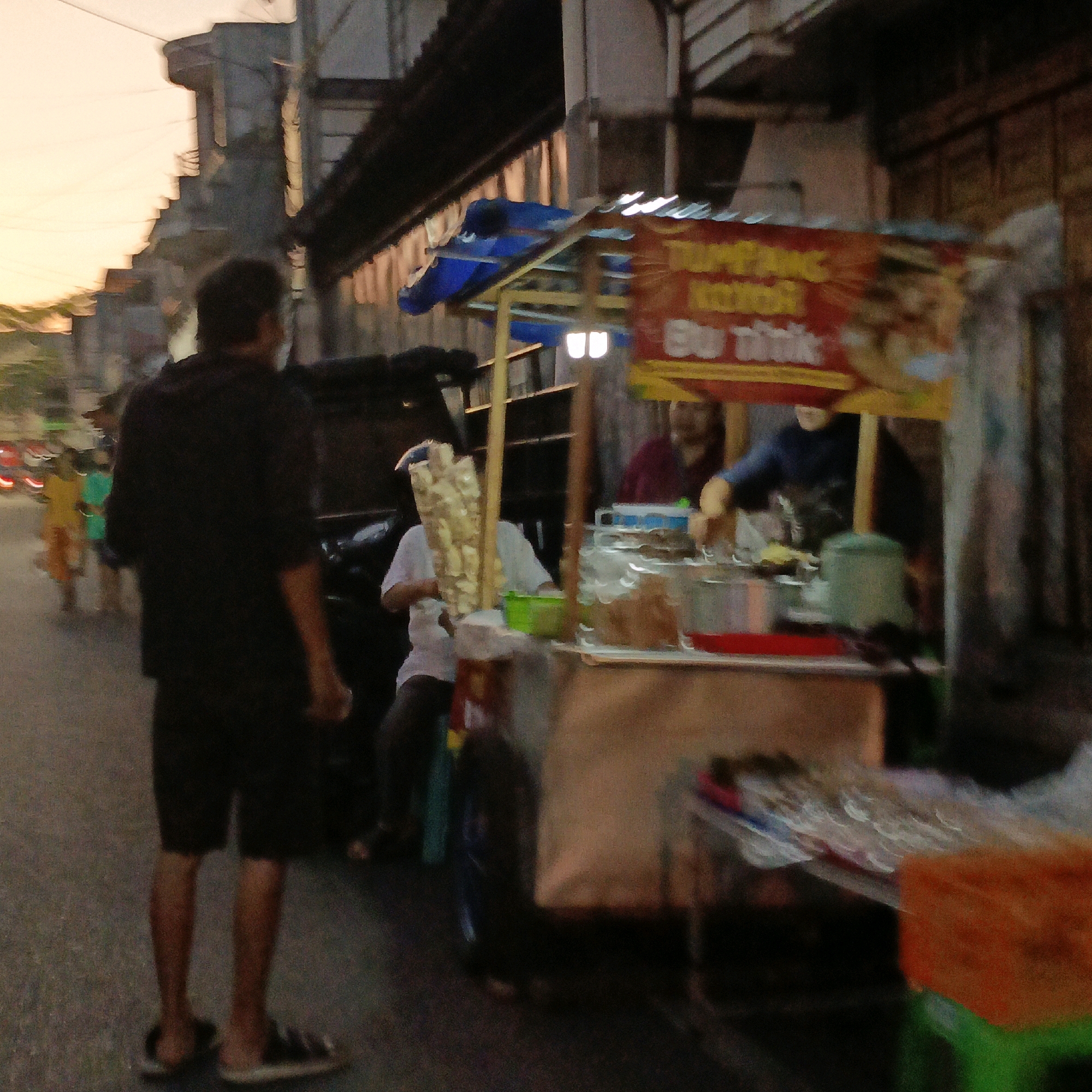 The image depicts a street scene in Salatiga, Indonesia, likely during dusk or early evening, indicated by the soft, warm lighting and the blurry motion suggestive of a low-light, potentially handheld, capture. The foreground is dominated by a street vendor's cart, laden with various food items and containers. A person in a dark shirt and shorts stands facing away from the camera, their back to the viewer, positioned on the left side of the frame. Behind them, on the street, are other individuals, some appearing as blurred figures in brightly colored clothing, suggesting movement. The cart itself has a blue tarp roof and a sign with red and yellow lettering, partially obscured. Visible on the cart are containers filled with food, possibly snacks or ingredients, and a green plastic bowl. The background shows buildings with a traditional architectural style, lining a street that appears to be a commercial area. The overall impression is a candid snapshot of everyday life in a bustling Indonesian city.