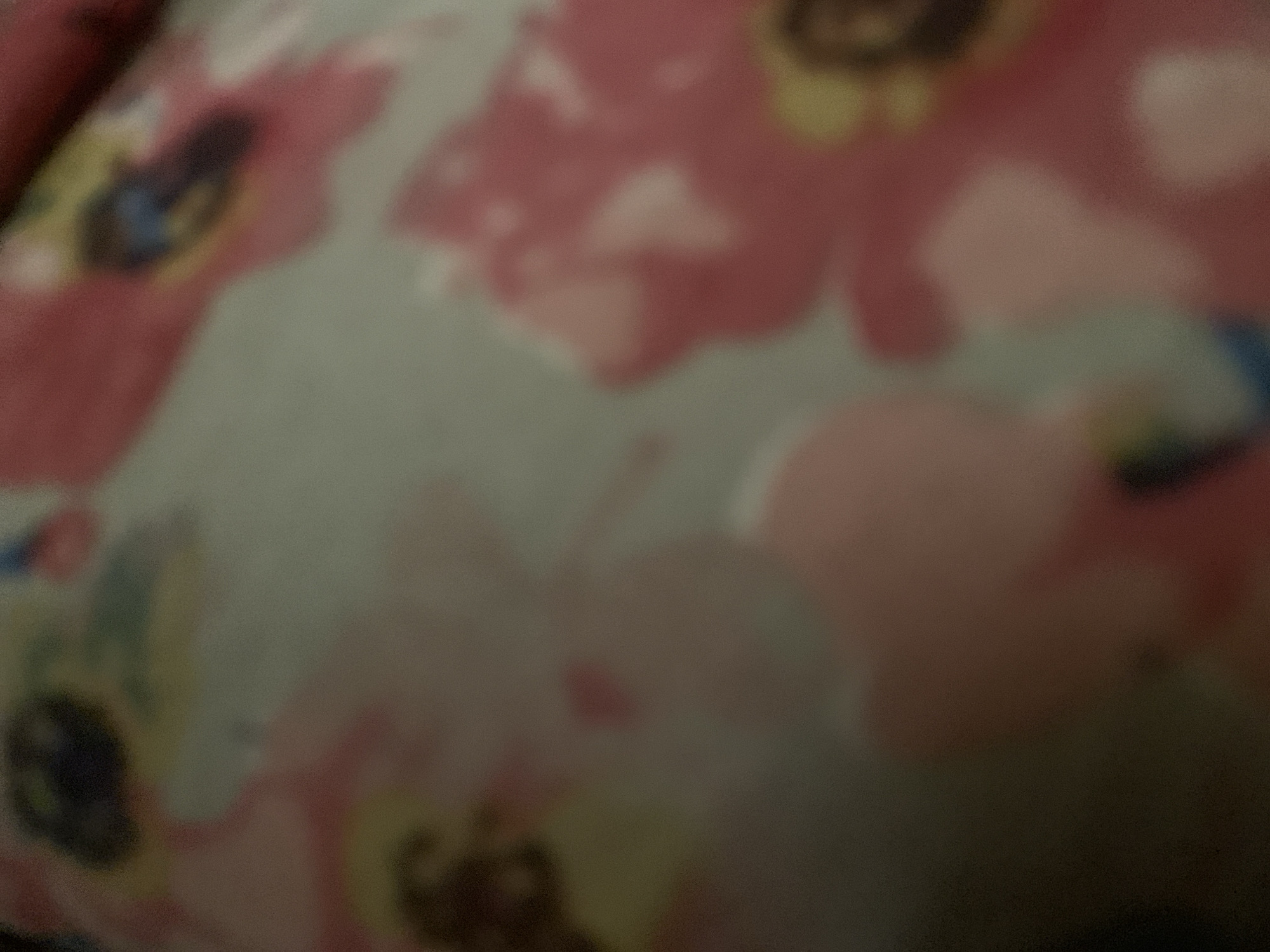 The image is a close-up, out-of-focus shot of a floral fabric. The fabric features large, abstract pink and red flowers with yellow centers, set against a muted green background. Some darker blue and purple elements are also visible within the floral patterns. The image lacks specific details related to Jalingo, Nigeria, as it is a still life of fabric with no discernible context of location or activity within the city.