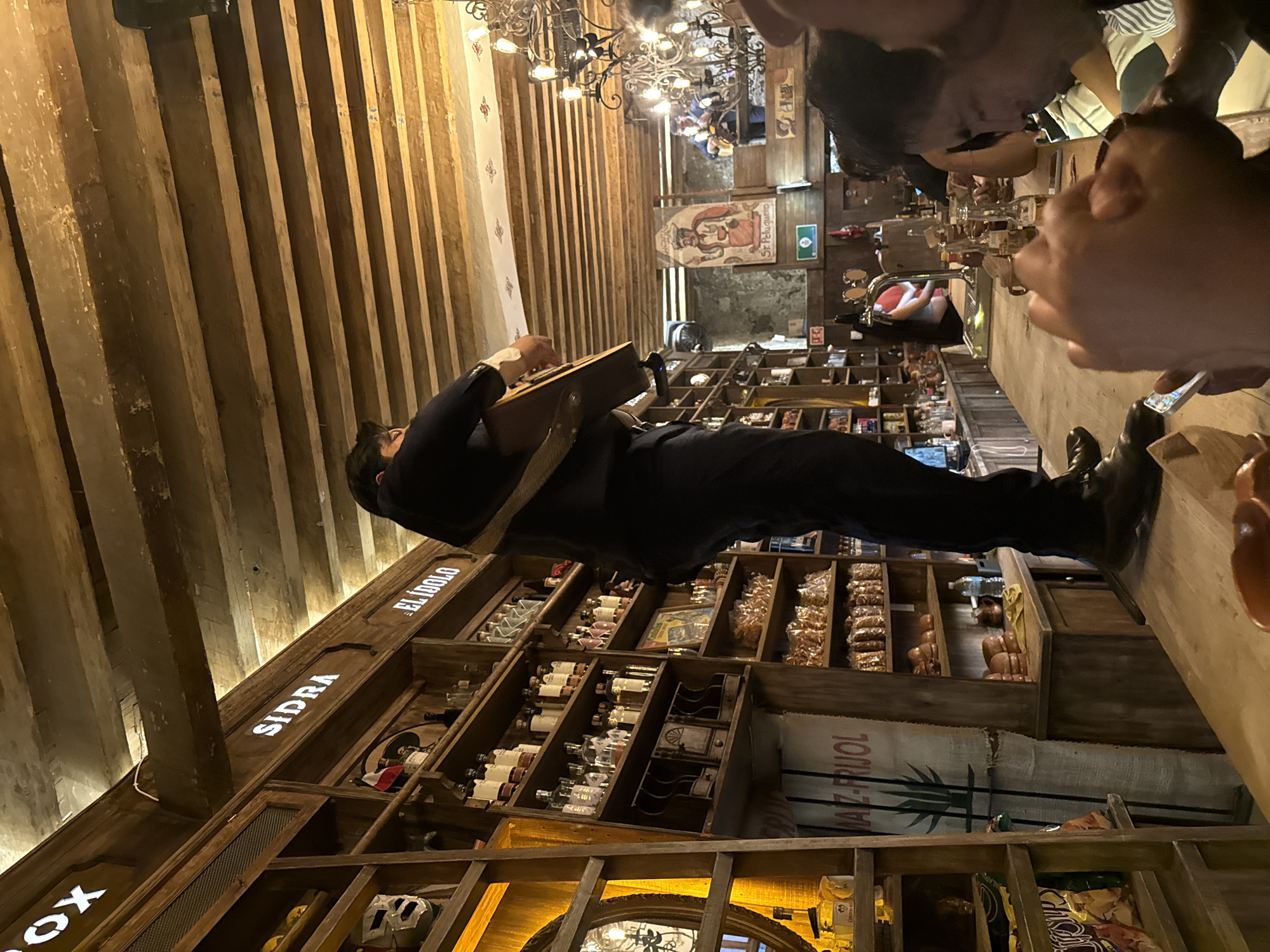 The image captures the interior of a bar or restaurant in Puebla, Mexico. The setting is characterized by rustic decor, featuring wooden beams on the ceiling and shelves stocked with various bottles, food items, and objects.
A man is playing a stringed instrument, likely a guitar or similar instrument, while standing. He is dressed in dark clothing. The presence of other people at the bar suggests a social atmosphere. There are some people at the bar using their phones.