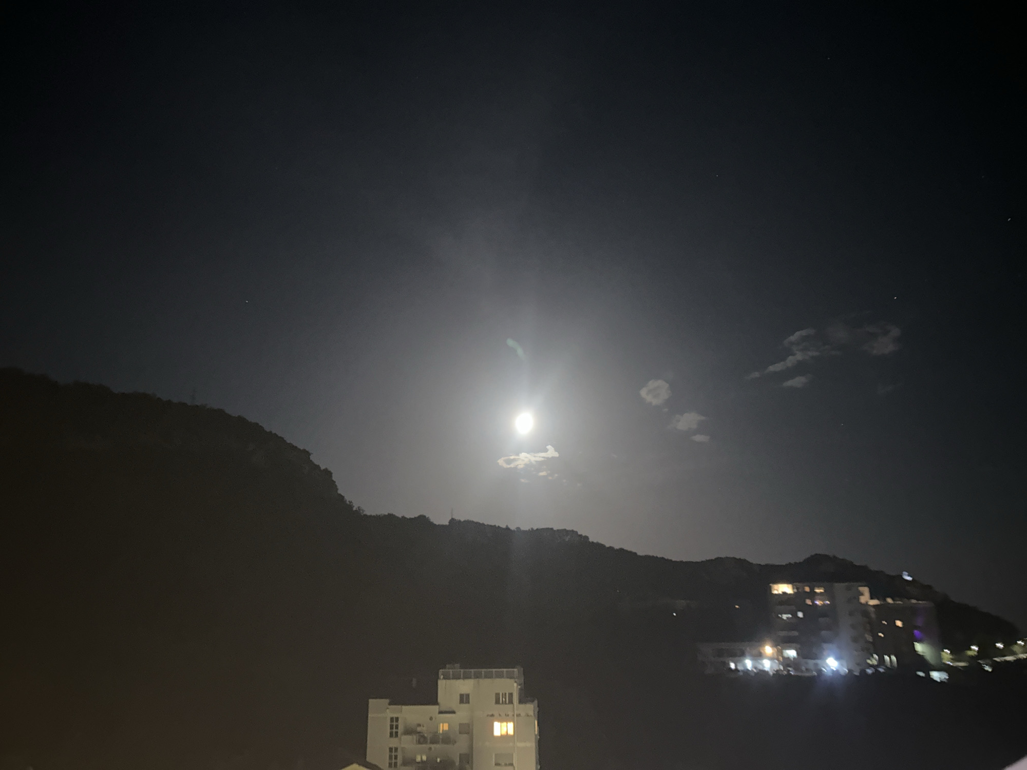 This is a nighttime photograph taken in Rafailovići, Montenegro. The scene depicts a mountain range silhouetted against a dark sky illuminated by a bright full moon. A few small clouds are visible near the moon. In the foreground, there are buildings with illuminated windows. One building, in the lower left of the frame, appears to be a multi-story apartment or hotel building. Further to the right, another larger building, possibly a resort or hotel complex, is visible with many illuminated windows and some external lighting. The overall impression is a serene, nocturnal landscape of a coastal town.