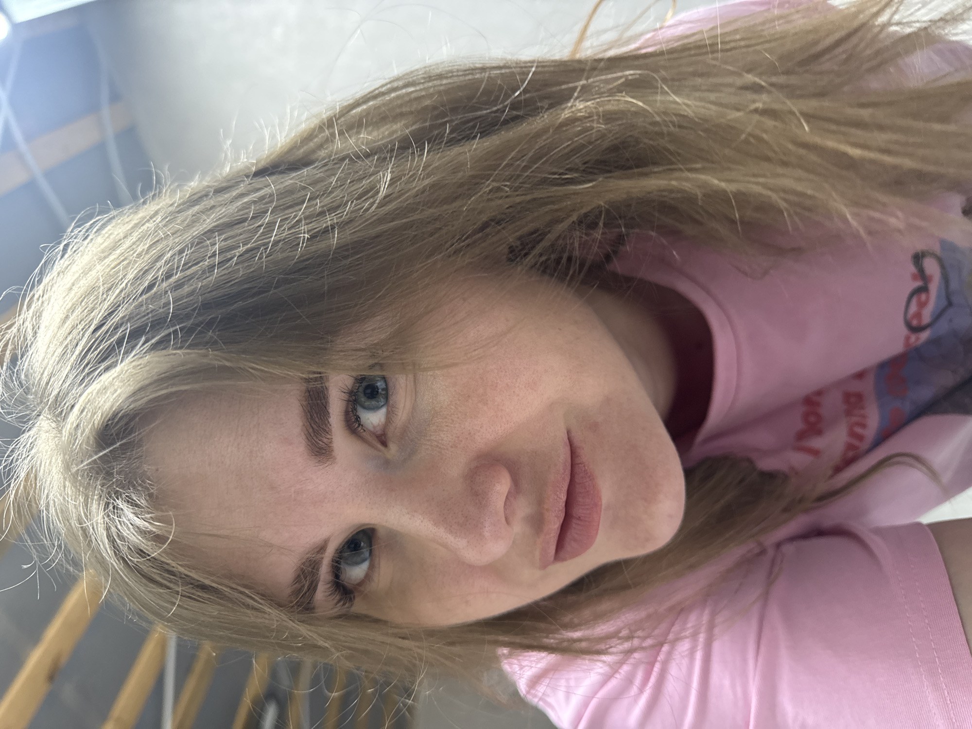 A woman with long, light brown hair and blue eyes is wearing a pink t-shirt. The photograph appears to be taken from a slightly above, tilted angle. The background is out of focus, but a light-colored wall and what looks like wooden stairs are visible. There is no specific indication of the location being Gornyi (Dvurechenskoe S/P), Russia.