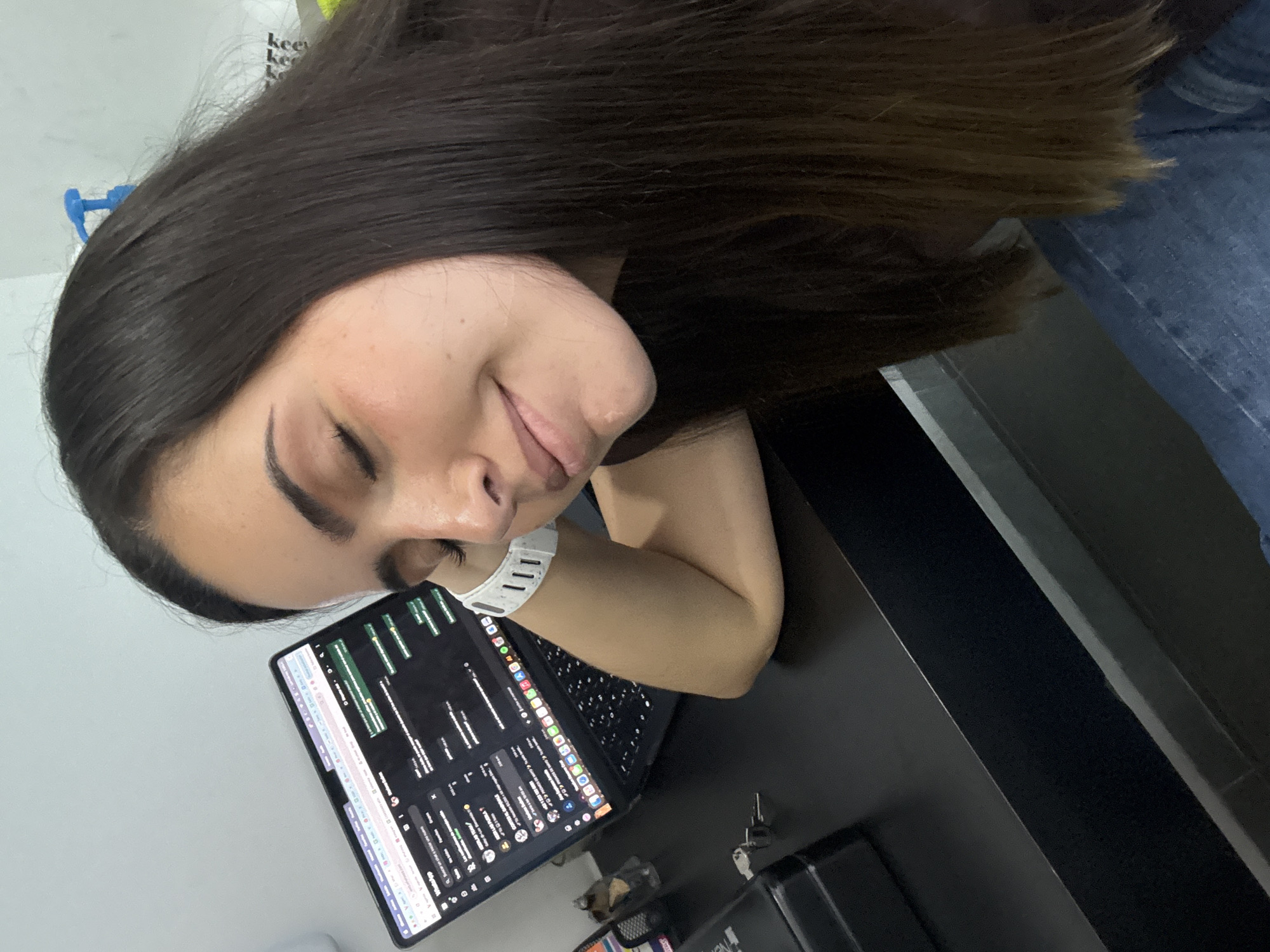 A woman with long dark hair is looking down with a slight smile. She is wearing a light-colored smartwatch on her left wrist. Her arm rests on a black desk. A tablet computer is open on the desk, displaying what appears to be a communications interface or a coding environment with numerous text-based windows and icons. There are keys and other small objects on the desk as well. The context suggests a person working in an office environment, but there is no specific indication of the location being San Bernardino Tlaxcalancingo, Mexico.
