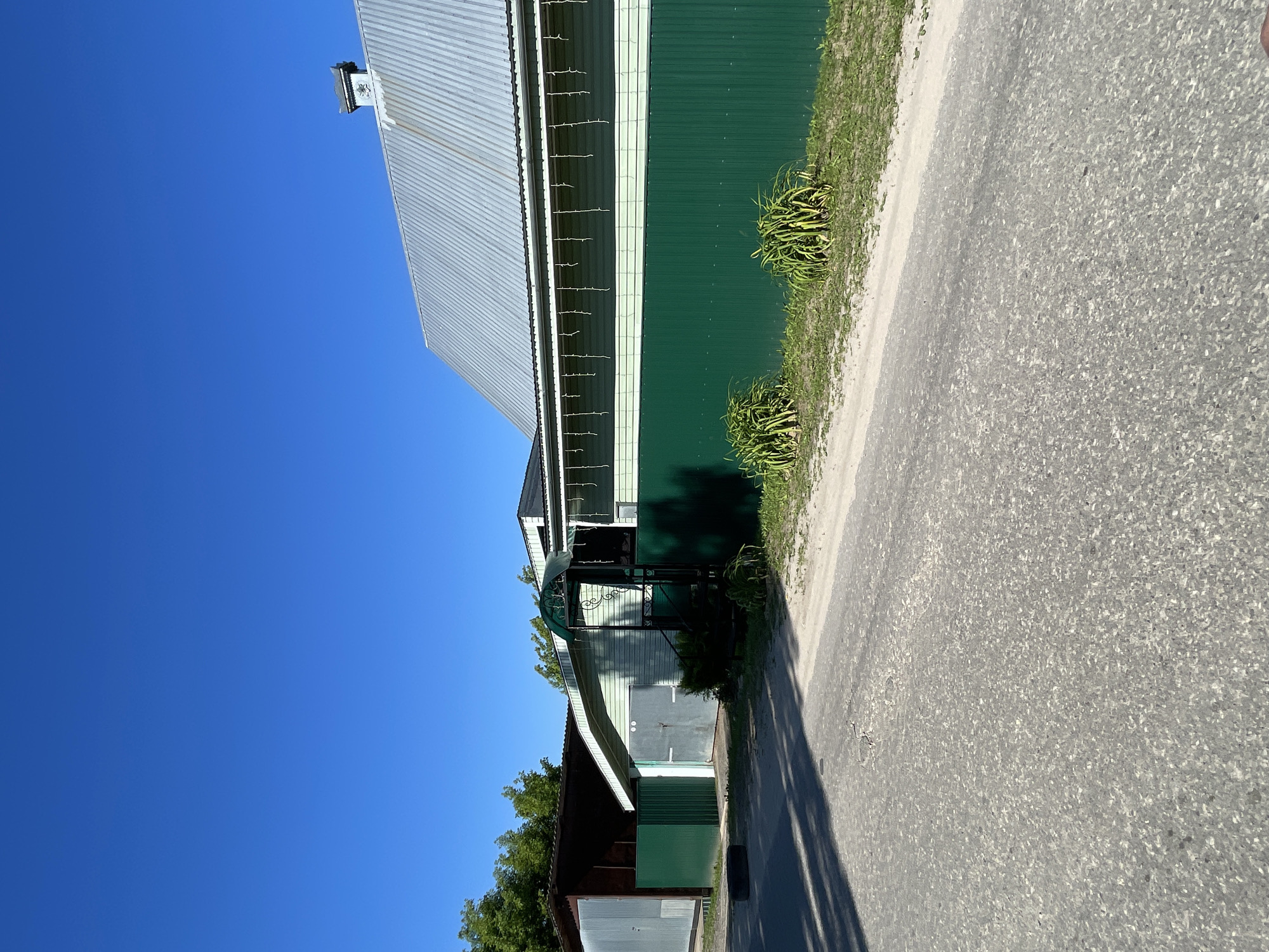 The image shows a residential property on a sunny day. A light blue sky dominates the upper portion of the frame. In the foreground, a grey asphalt road runs diagonally from the lower left to the middle right. The road has some minor cracks and imperfections. To the right of the road, there is a strip of sandy ground with green grass and two clusters of leafy green plants.
Beyond the sandy area, a dark green metal fence encloses the property. Behind the fence, a light green building with white trim and a corrugated metal roof is visible. Icicle-shaped lights are strung along the top of the light green siding. To the left of this building, there is a darker building with a sloped roof and a large grey metal garage door. Next to that, another dark building, possibly a garage, has a dark brown exterior and a lighter colored metal door. A green metal gate is visible in front of this structure. A black tire lies on the ground near the green gate. A chimney with a decorative top is visible on the metal roof of the main building. The scene suggests a quiet, possibly rural or suburban, setting in Красные Баки, Russia.