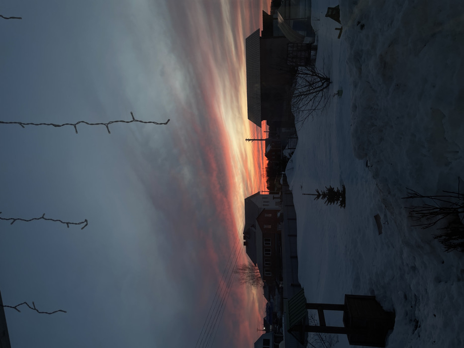 The scene depicts a winter landscape at dusk in Olkhovka, Russia. The sky transitions from a deep blue-grey at the top to vibrant reds, oranges, and yellows along the horizon, streaked with clouds. In the immediate foreground, three dark, unlit strands of string lights hang vertically from an unseen structure above. The ground is uniformly covered in snow, forming drifts and uneven terrain. To the left, a wooden well structure with a dark-pitched roof is partially visible, while a small, snow-covered evergreen tree stands centrally. Bare tree branches protrude from the snow in the lower portion of the frame. The midground features a two-story house with dark walls and a dark roof on the left, and a larger, dark-silhouetted building with a lighter, possibly metal roof on the right. A utility pole with power lines rises from the left, silhouetted against the bright horizon. Additional structures and bare trees are visible in the distant background. No individuals are present.