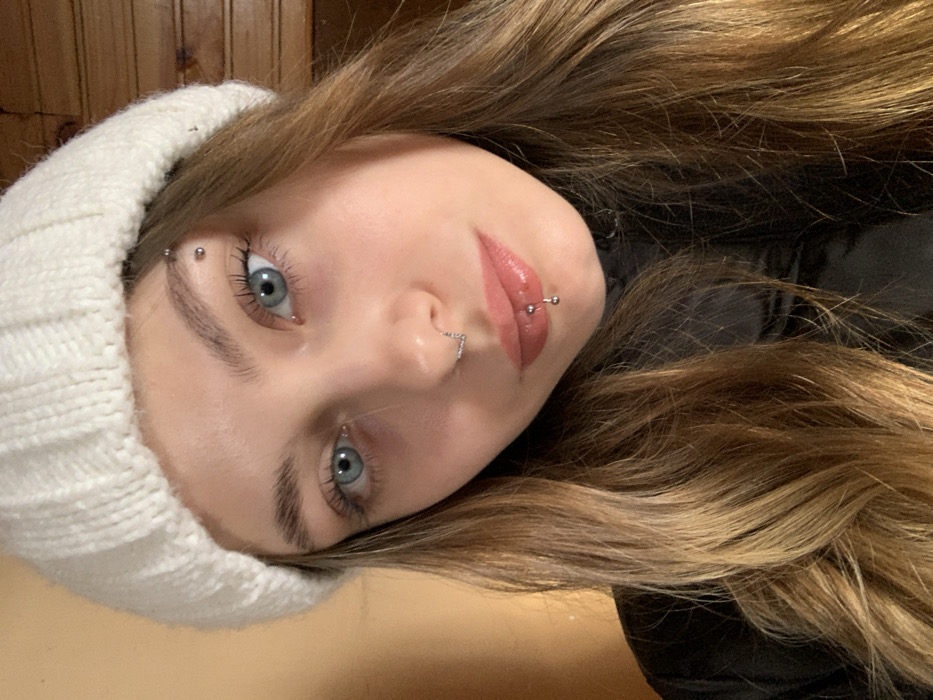 A young woman with light blue eyes gazes at the camera. She wears a white knitted beanie, and her long, highlighted brown hair frames her face. Her eyebrows are pierced with a small barbell, and she has a nose piercing. Her lips are glossy and pierced with a labret barbell. The background is blurred, showing a wooden-paneled wall and a glimpse of a black garment. The photo is taken from a slightly tilted angle.
