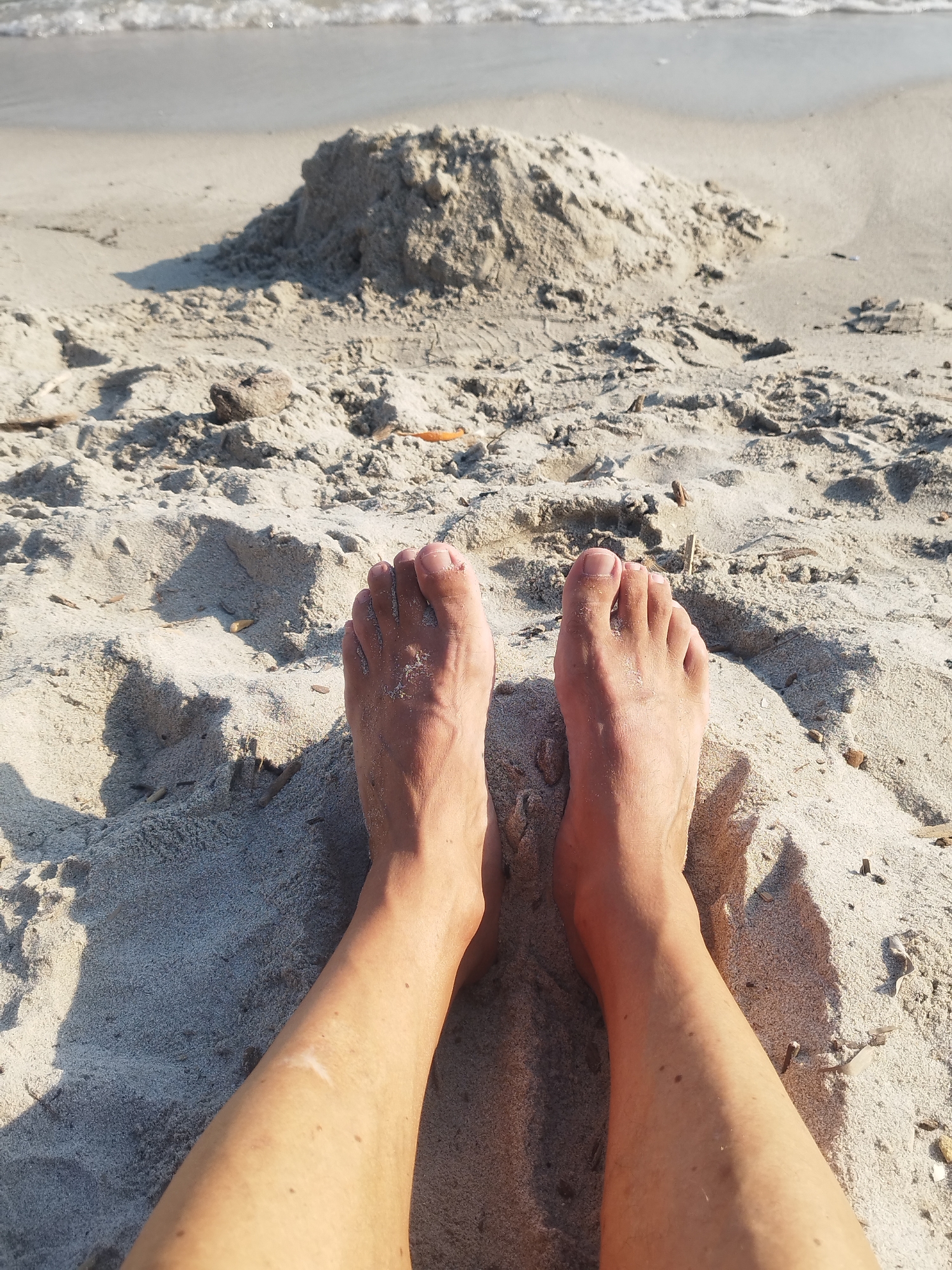 This is a close-up, eye-level shot of a person's bare feet partially buried in the sand on a beach. The sand is light-colored and textured, with small pebbles and pieces of debris scattered throughout. In the background, the ocean is visible with gentle waves lapping at the shore. The lighting suggests it is daytime, with the sun casting shadows on the sand. The image does not contain any identifiable landmarks or specific features that would place it in Antibes, France.