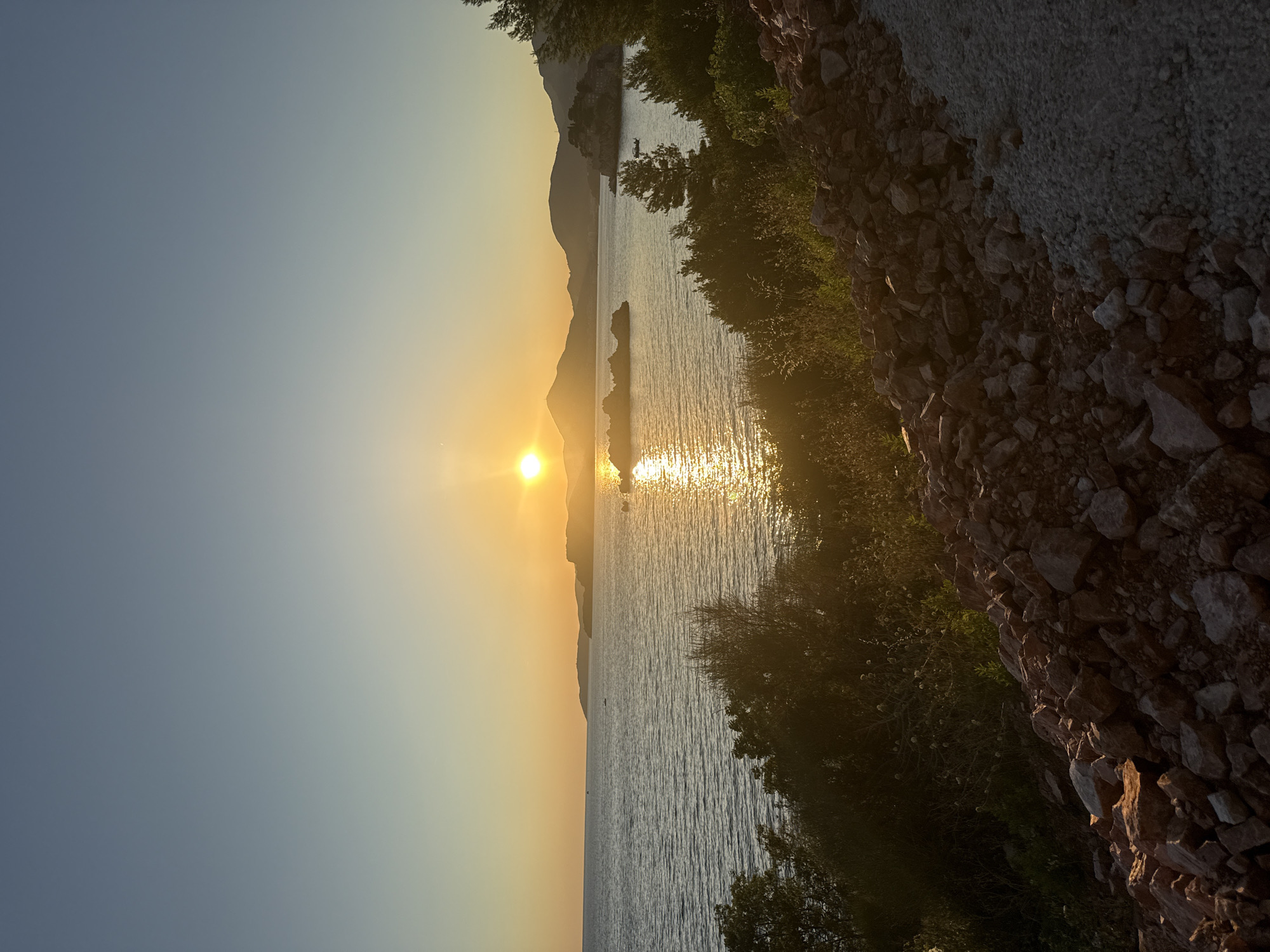 A vertical, eye-level, wide shot captures a serene sunset over the Adriatic Sea, viewed from a rocky incline near Sveti Stefan, Montenegro. The sun, a bright, warm orb, is positioned slightly right of center in the upper half of the frame, casting a golden reflection across the water. The sky transitions from a pale blue at the top to a soft yellow and orange near the horizon. Silhouetted against the setting sun are rolling hills and mountains, with a small, fortified island visible in the middle distance, a characteristic of Sveti Stefan. The water is calm, with gentle ripples reflecting the light. In the foreground, the incline is covered with rough, reddish-brown rocks and sparse vegetation. A path of light-colored gravel is visible on the right side of the foreground. The overall atmosphere is tranquil and picturesque. There are no people or animals visible in the image.