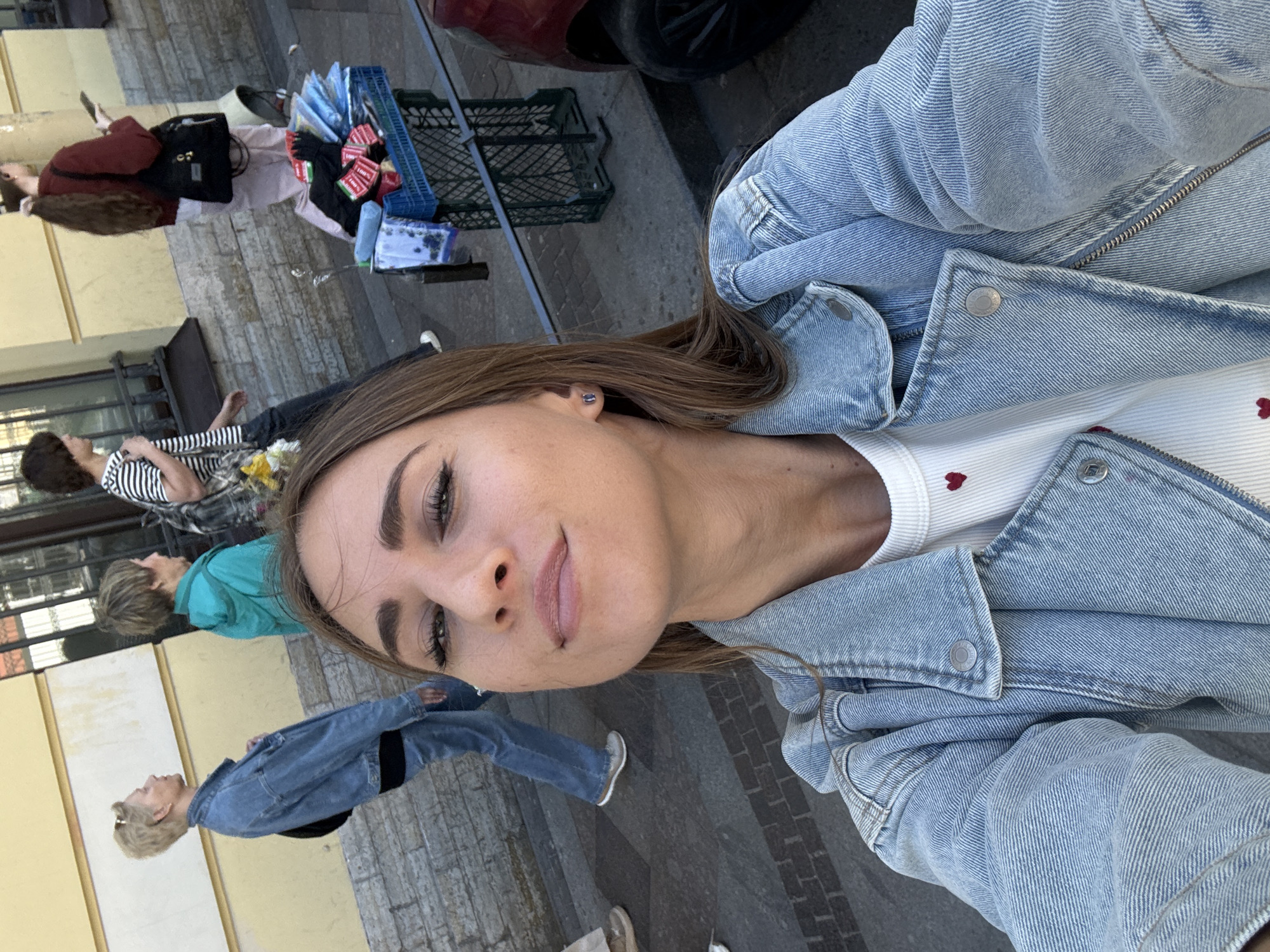 In Saint Petersburg, Russia, a woman with long brown hair, wearing a denim jacket over a white top with red hearts, takes a selfie outdoors on a sidewalk with a yellow building in the background. Several people are walking past. To the right, a blue plastic crate displays various items, possibly for sale, including clothing and socks. A red car is partially visible on the right edge of the frame. The ground is paved with cobblestones and stone tiles.