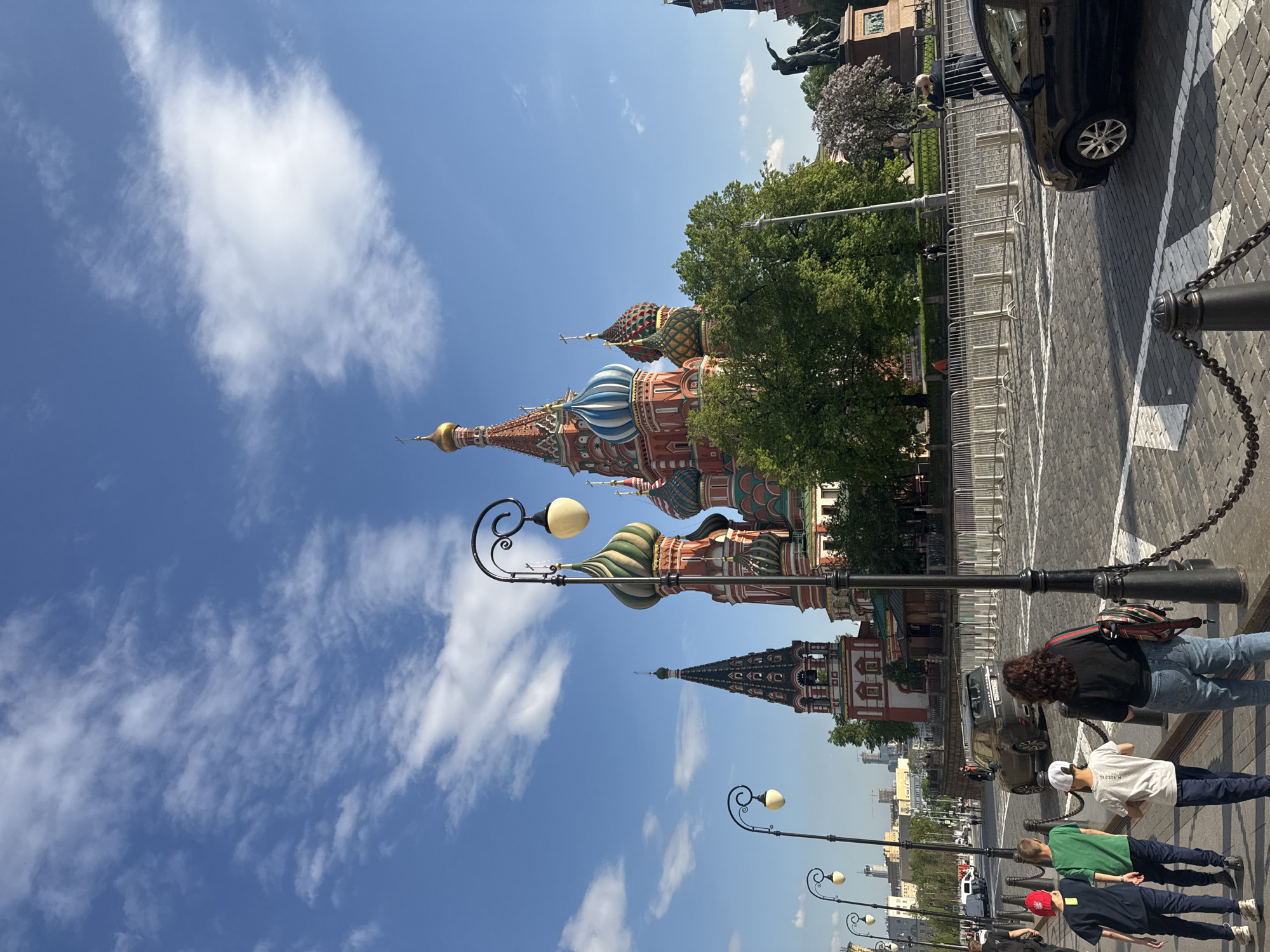 The image depicts a street scene in Moscow, Russia, showcasing the iconic St. Basil's Cathedral. The cathedral is visible with its distinct, multi-colored domes. The sky is blue with scattered white clouds. Several people are walking along the street. Street lamps line the pavement. A dark-colored car is parked near a metal fence. The street is paved with cobblestones. A monument is visible in the background, behind the trees, in front of St. Basil's Cathedral.