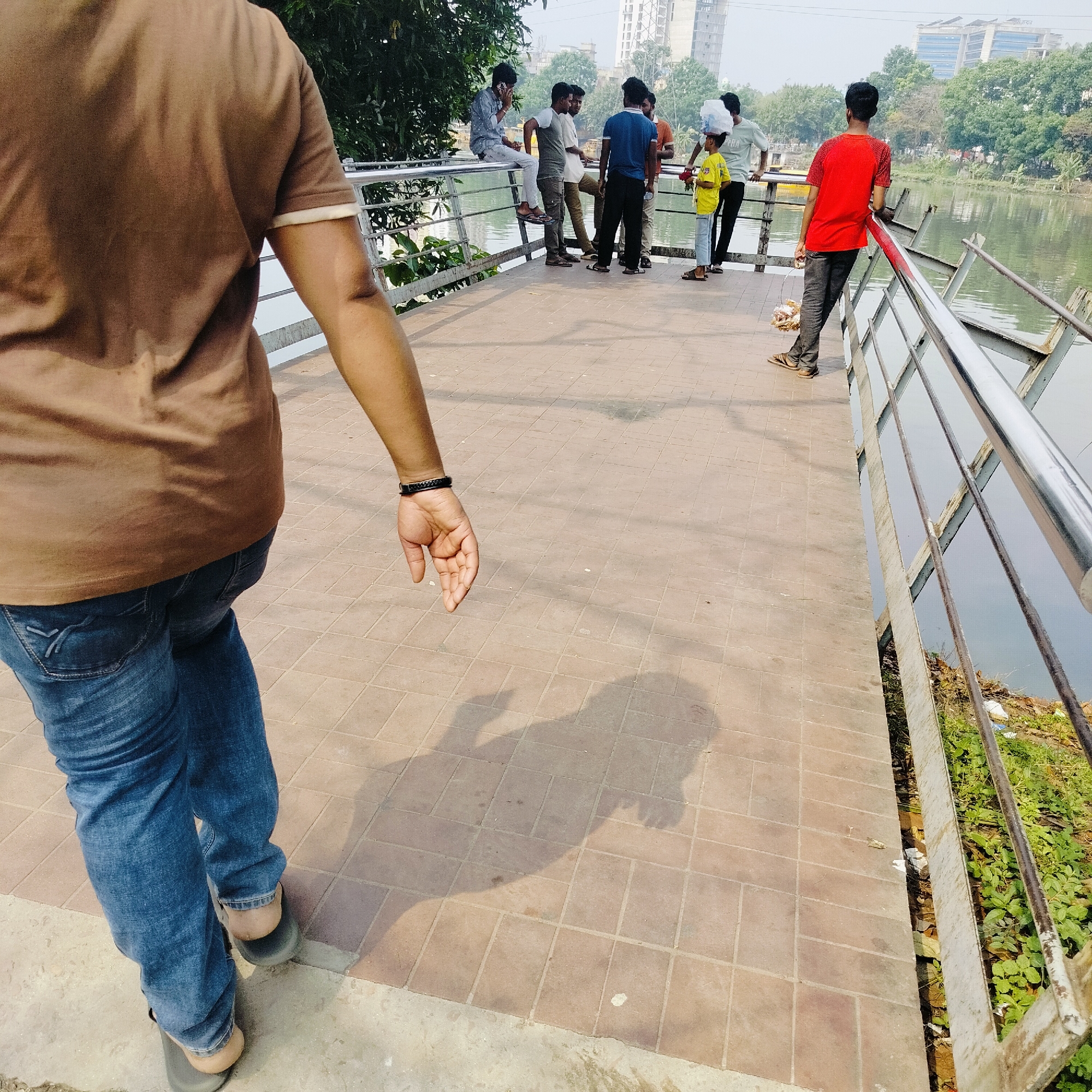 The image depicts a group of people on a walkway in Dhaka, Bangladesh. In the foreground, a person wearing a brown shirt and jeans is walking towards the group. A shadow of a person is cast on the tiled walkway. In the background, several individuals are standing and sitting near a railing overlooking a body of water. The environment includes vegetation along the water's edge and buildings in the distance. The walkway appears to be constructed of brown tiles. The water appears to be a lake or pond.