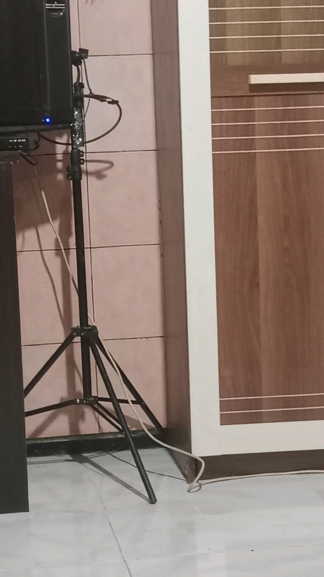 An indoor scene is depicted, likely a room, with a tiled wall in the background and a tiled floor in the foreground. On the left, a black speaker is partially visible, mounted on a black tripod stand. A black cable extends from the speaker down the stand. To the right of the speaker and tripod, a large wooden wardrobe or cabinet is present, with a vertical white trim and horizontal lines across its surface. A white cable runs across the floor from the vicinity of the tripod towards the wardrobe. The lighting suggests a domestic environment. There are no people visible in the image, nor are there any explicit indications of the specific location within Kebonagung, Indonesia.