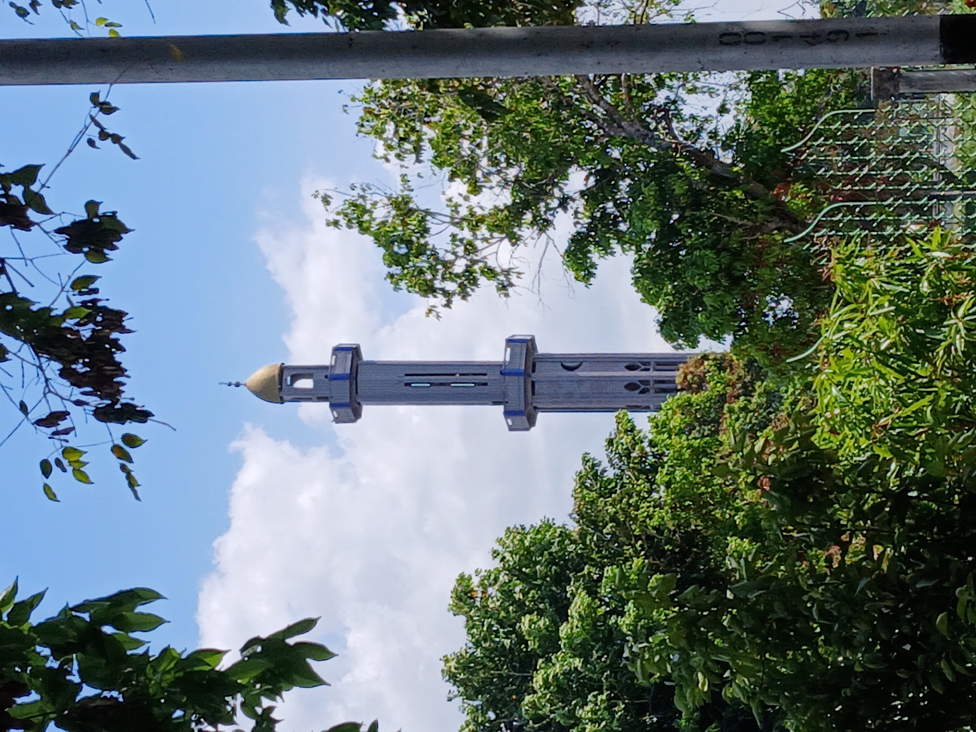 The image shows a tall, grey mosque minaret with a golden dome, framed by green trees and a blue sky with white clouds. A grey metal bar is visible in the upper portion of the image, partially obscuring the view. The location is Ciudad Guayana, Venezuela.