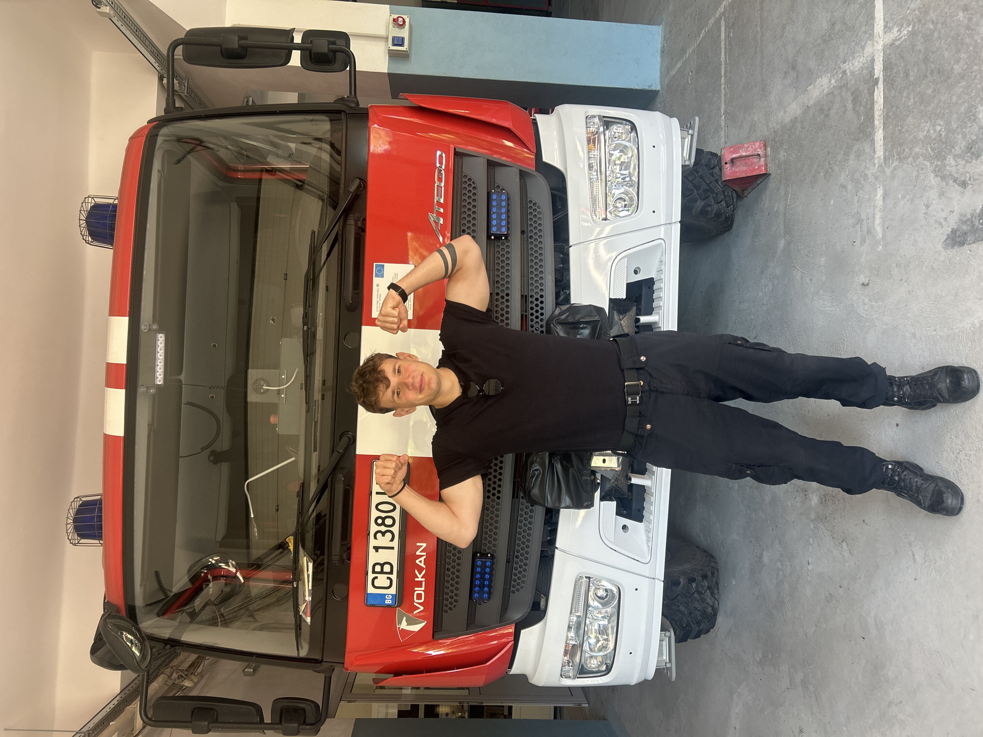 A young man in black clothing and boots poses in front of a fire engine in what appears to be a garage or station in Sofia, Bulgaria. The fire engine is red and white and features a Bulgarian license plate with the code "CB 1380 BG," which is indicative of Sofia. The man is standing with his back against the engine's front, flexing his arms. The environment is a garage or station, with the fire engine as the primary focus.