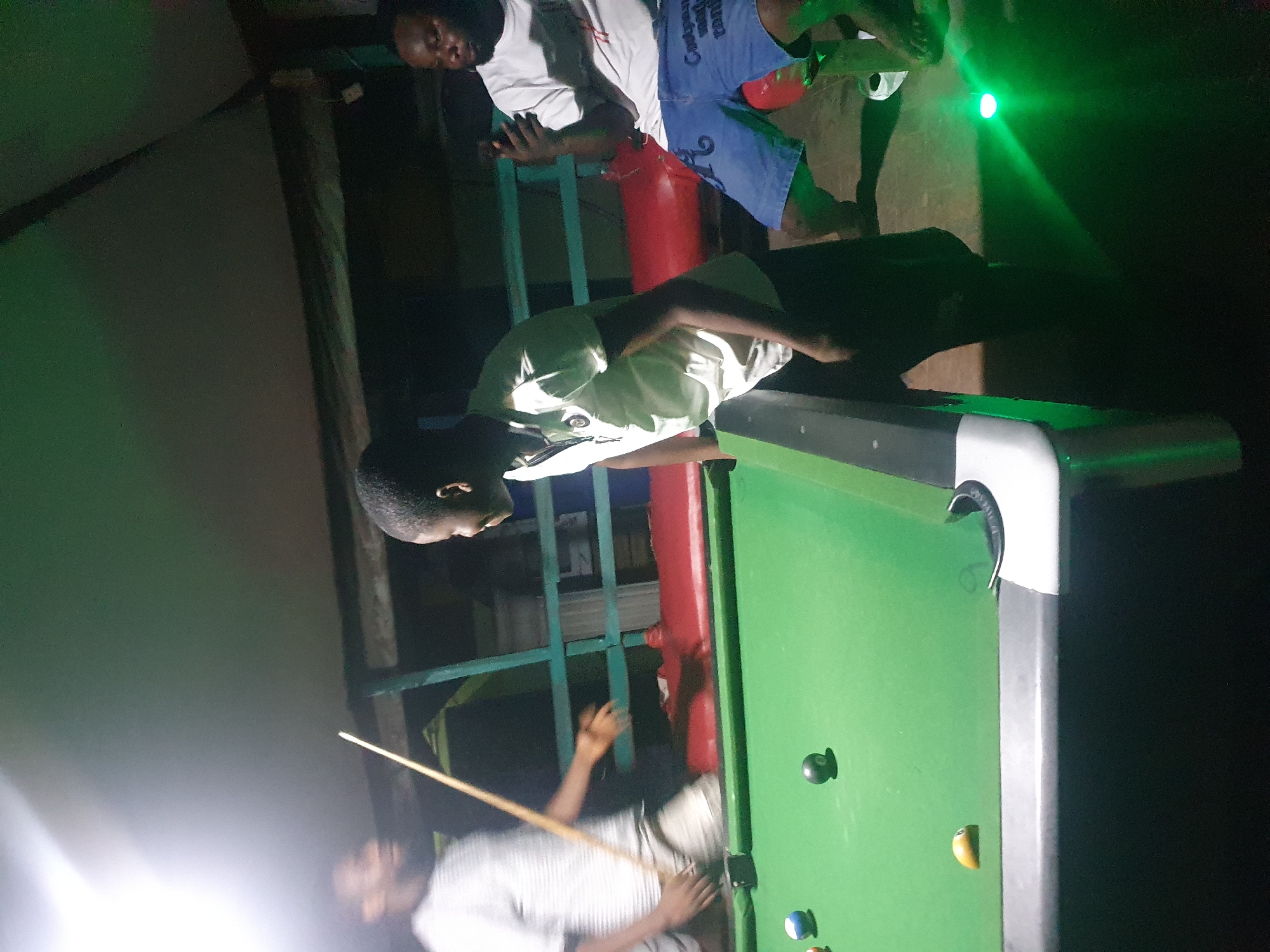 In Lokoja, Nigeria, at night, three men are engaged in a game of pool. The primary focus is on a pool table with a green felt surface, dotted with several billiard balls, including a black 8-ball and a yellow ball. Two individuals are actively involved with the table: one man, wearing a white striped t-shirt and light-colored shorts, is positioned to strike a ball with a cue stick. Another man, to his right, is observing the game. The third man is seated to the far right, holding a mobile phone and looking down at it. The environment appears to be an outdoor or semi-outdoor setting, possibly a bar or recreational area, characterized by low lighting, wooden structures, and a green light source emanating from the lower right of the frame.