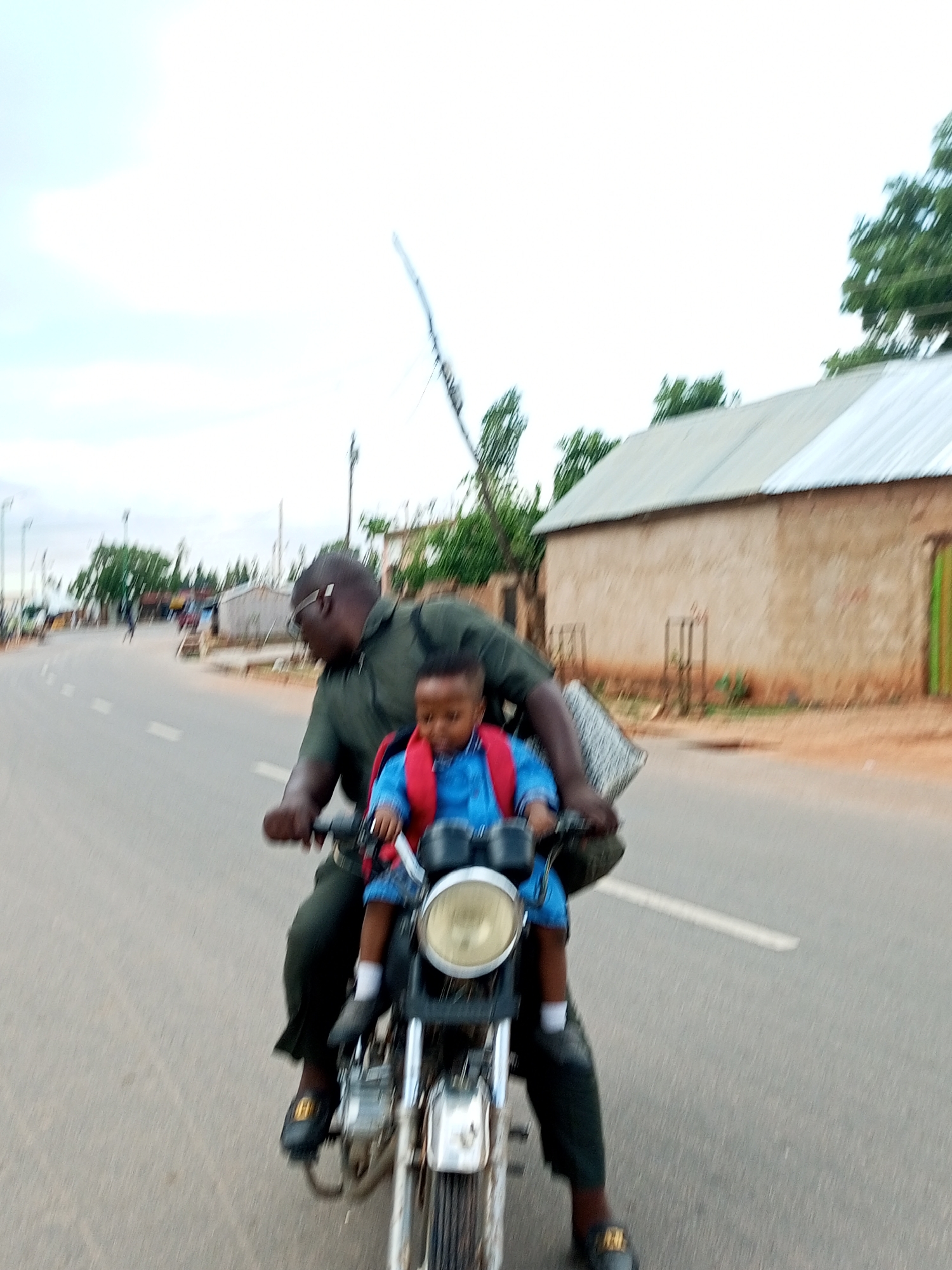 In Tempure, Nigeria, a man in a dark green shirt and dark pants, wearing glasses, is riding a motorcycle. He is carrying a young child in front of him who is wearing a blue jacket with a red backpack. The child has white socks and dark shoes. The motorcycle has a visible headlight and appears to be moving on a paved road. In the background, there are buildings with corrugated metal roofs and trees. The sky is overcast.