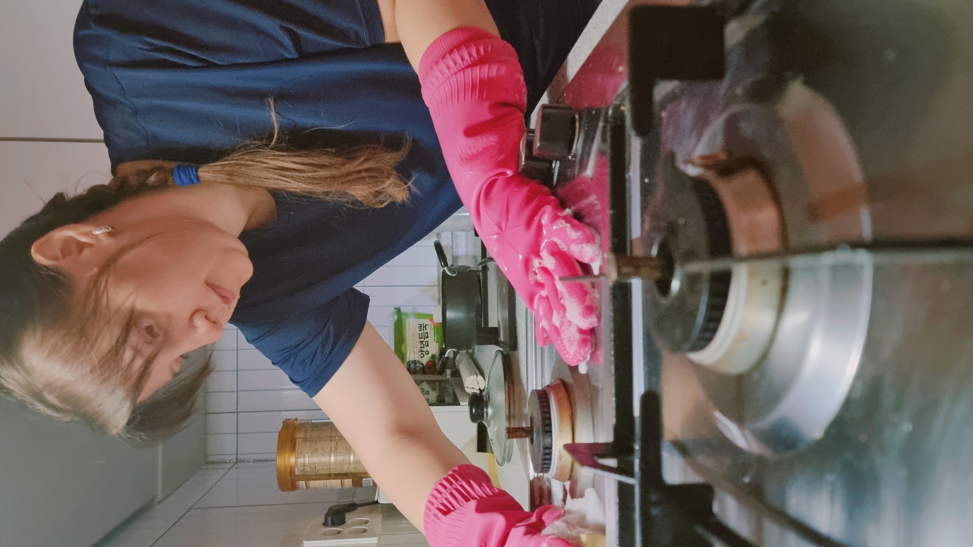In Dunnae-myeon, South Korea, a woman with light brown hair pulled back with a blue hair tie is cleaning a gas stovetop. She is wearing a dark blue t-shirt and pink rubber gloves. Her hands are covered in soap suds as she scrubs the stovetop. In the background, there is a jar, a box of food, a pot and lid, and white tiled walls.