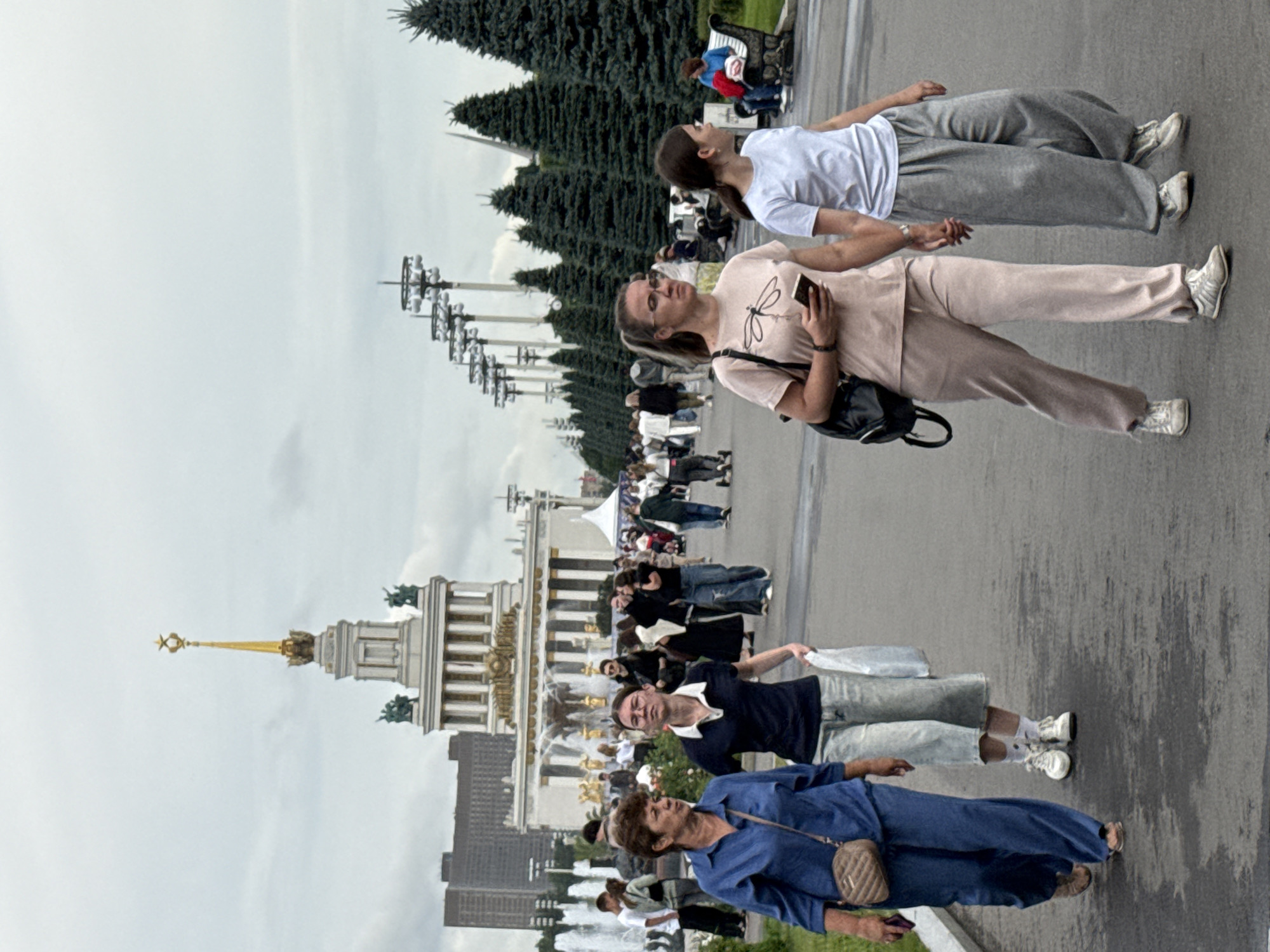In Moscow, Russia, at the VDNKh exhibition center, a group of people walk along a paved path under a cloudy sky. In the foreground, two women walk side-by-side, one in a blue tracksuit and the other in a black shirt and light-colored cropped pants. Behind them, two other women walk together, one in a light pink tracksuit, holding a mobile phone, and the other in a white t-shirt and grey sweatpants, holding hands. In the background, a large ornate building with a golden spire and statues stands prominently, with a row of tall fir trees lining the path and a distant cityscape visible. Other individuals and groups are scattered throughout the scene, some near fountains and others sitting on benches.