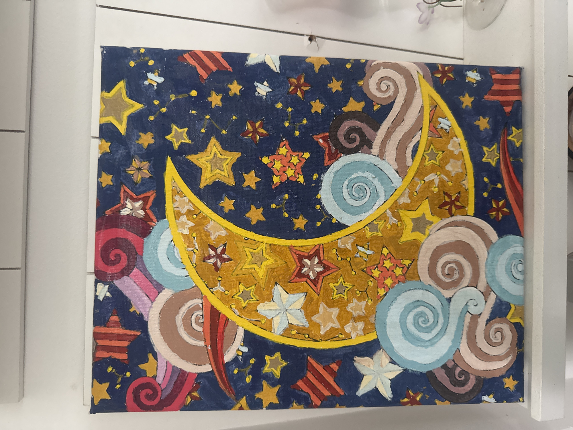 The image is a close-up shot of a painting. The painting depicts a blue night sky filled with various sized yellow and orange stars. A large, crescent moon fills the center of the painting, outlined in a bright yellow. The inside of the moon is filled with more stars and swirls. Around the moon are swirls of brown, light blue, and reddish-purple, possibly representing clouds or celestial bodies. The painting appears to be hanging on a wall. The location is not discernible from the image, and there is no indication of its location in Plovdiv, Bulgaria.