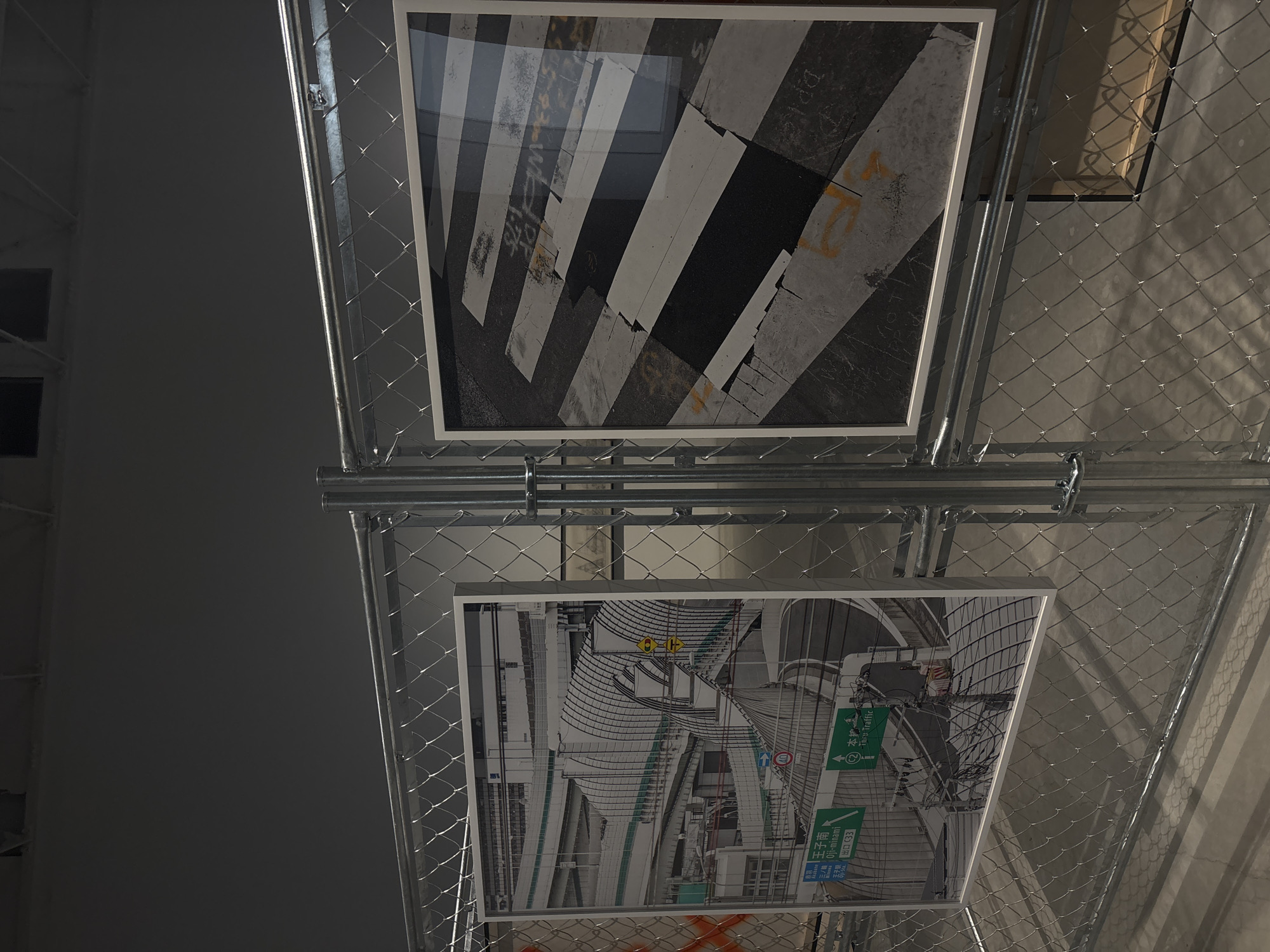 The image displays an indoor art installation featuring two framed photographs mounted on a grey metal mesh structure, resembling a large industrial fence. The background is a plain, dark grey wall on the left, transitioning to lighter concrete or ceiling elements on the right.
The top framed photograph presents a stark, graphic view of an urban crosswalk. Bold, weathered black and white stripes run diagonally across an asphalt road surface. Faint yellow painted markings and faded Japanese characters are subtly visible on the asphalt, adding to the authentic urban feel. The glossy surface of the frame reflects overhead light.
Below, the second framed photograph captures a complex urban landscape, likely an elevated highway or a multi-level road intersection. The scene is dominated by concrete ramps, supporting pillars, and a dense network of overhead wires, creating a dynamic architectural composition. Prominent green road signs feature white Japanese text with English transliterations, including "↑ 本線 Go Straight," "C2 環状線" (Loop Line C2), "王子南 Ōjimani," and "出口 C33" (Exit C33). A yellow diamond sign with an "H" symbol and a blue circular sign with a red diagonal line are also visible.
The photographs are secured within the sturdy, industrial-style mesh and metal piping framework. The visible Japanese text and specific road signs confirm the location as an urban area in Japan, consistent with the given context of Kanazawa. The display has a contemporary, perhaps minimalist, aesthetic. No people are present in the image.