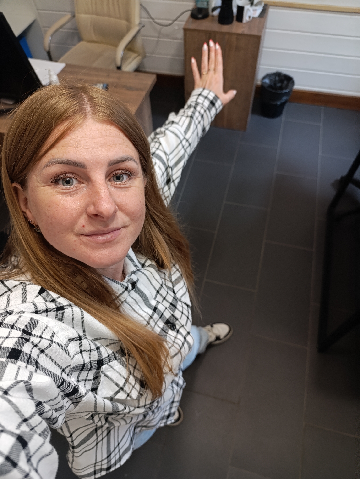 A woman with long, reddish-brown hair and blue eyes is taking a selfie in what appears to be an office or work environment. She is wearing a black and white plaid shirt and blue jeans. Her right hand is raised, with painted red fingernails, and she is wearing a ring on her right ring finger. She is standing on a dark gray tiled floor. In the background, there is a desk, an office chair, a wooden cabinet, and a black trash can. The lighting suggests an indoor setting with overhead lights.