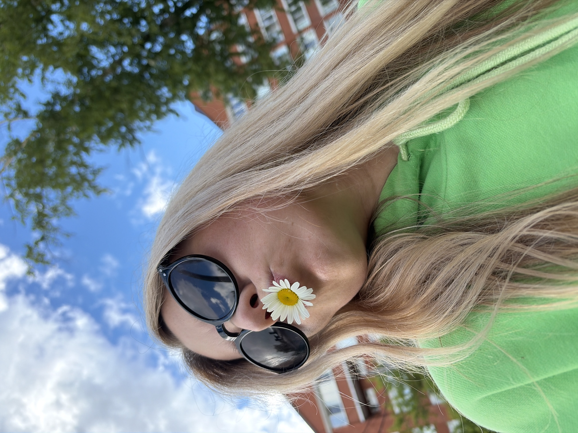 This close-up portrait image captures a person outdoors in Chaikovsky, Russia, during what appears to be a sunny day. The subject has long, flowing blonde hair that frames their face and extends into the foreground. They are wearing black sunglasses and a bright green hooded sweatshirt. A notable detail is a small white daisy with a yellow center, playfully placed on the bridge of their nose. The perspective is from a low angle, looking upwards. In the background, a clear blue sky with scattered white clouds is visible, along with blurred green tree foliage and a portion of a red-brick building with white window frames, indicating an urban or park setting. The image conveys a lighthearted and natural mood. No visible text is present.