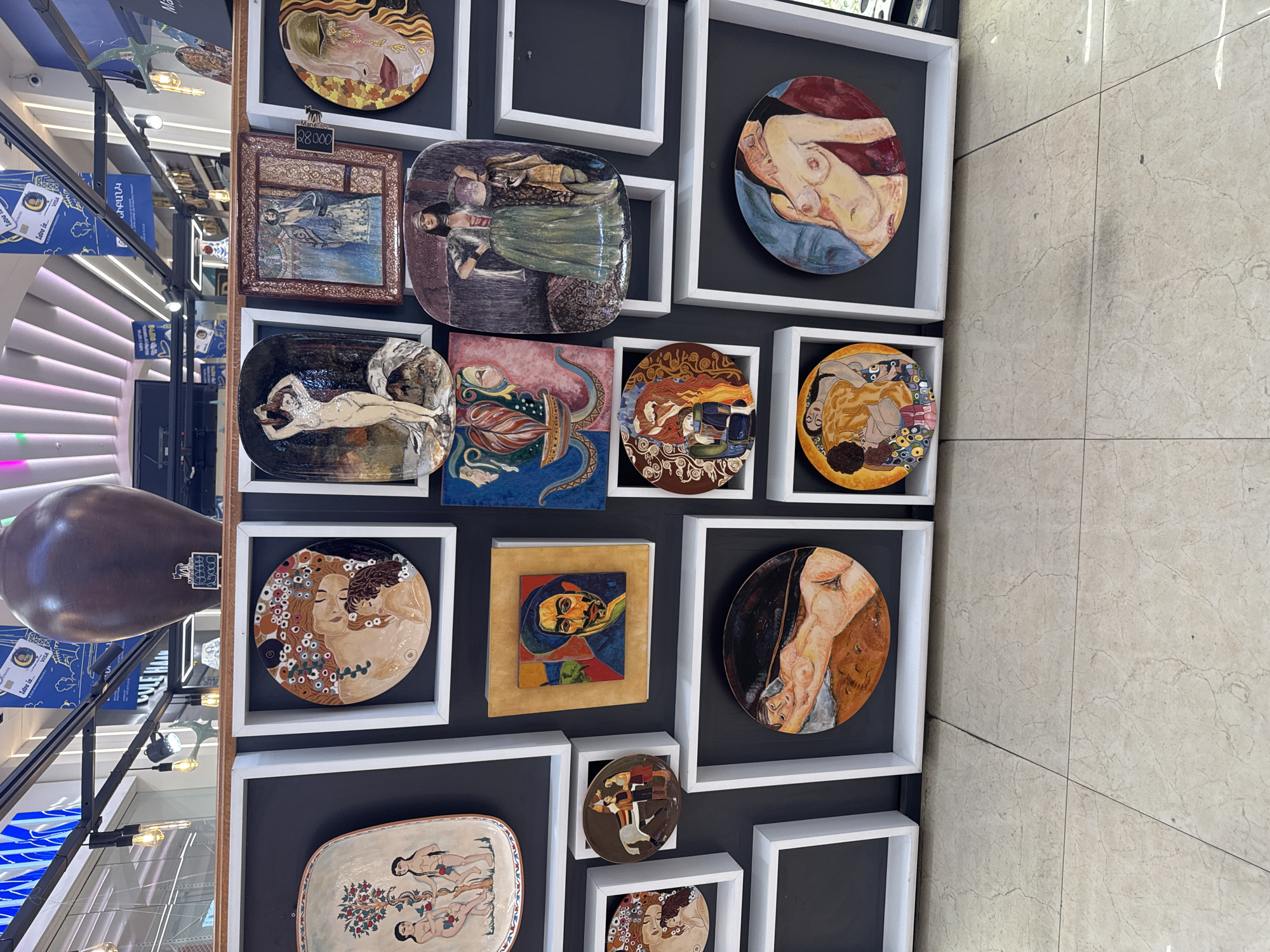 The image displays a collection of artworks, likely ceramic plates or tiles, mounted on a dark wall with white rectangular frames. The artworks feature various figurative paintings, including nudes, portraits, and mythological scenes. The style of the artwork appears to be a mix of classical and modern influences. In the background, a large, dark brown vase is visible, along with elements of a store interior, such as lighting fixtures and signage. One sign in the background is in Armenian script, suggesting the location is in Armenia. The overall impression is of an art exhibition or display. The floor in the foreground is tiled with a light-colored marble pattern.