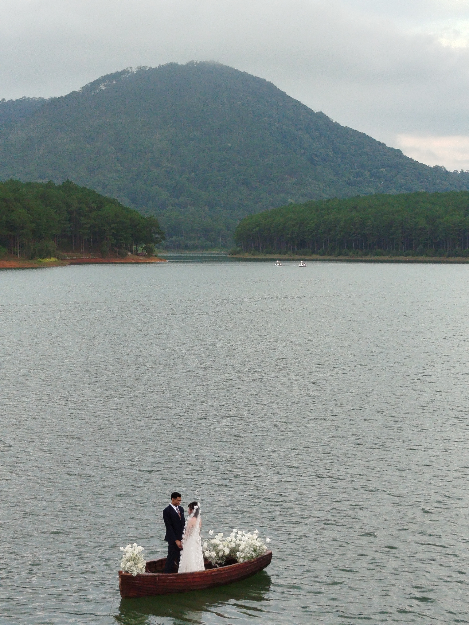 A wedding couple stands in a wooden rowboat on a calm lake, surrounded by white floral arrangements. The groom, wearing a dark suit and tie, faces the bride, who is in a white wedding dress and veil. The boat is positioned in the foreground, with the vast expanse of the lake stretching out behind it.
In the mid-ground, two small, white swan-shaped paddle boats are visible on the water. The background features a dense, green forest lining the far shore, leading up to a large, tree-covered mountain under a hazy, overcast sky. The scene suggests a serene and romantic wedding photoshoot.
The lighting indicates it is likely daytime, possibly late afternoon or early morning, given the diffused light and cloudy conditions. The location appears to be a picturesque natural landscape, consistent with the scenery of Da Lat, Vietnam, known for its lakes, forests, and mountains. No visible text is present in the image.