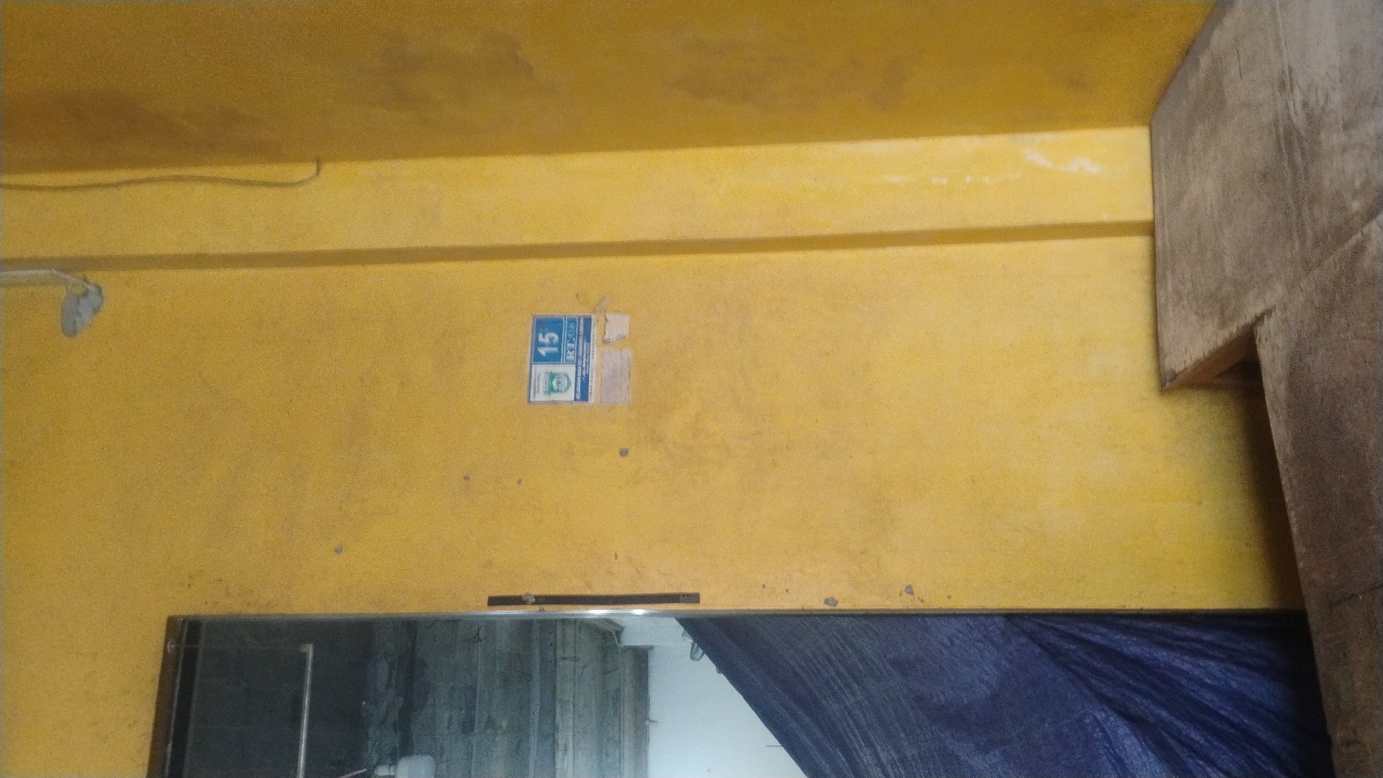 The image captures an interior scene with yellow-painted walls and a corner of a wooden structure. A blue tarp is visible on the left, partially obscuring an entryway. A sticker with the number "15" and some text in Indonesian is affixed to the wall. The overall impression is of a simple, possibly utilitarian, indoor space.