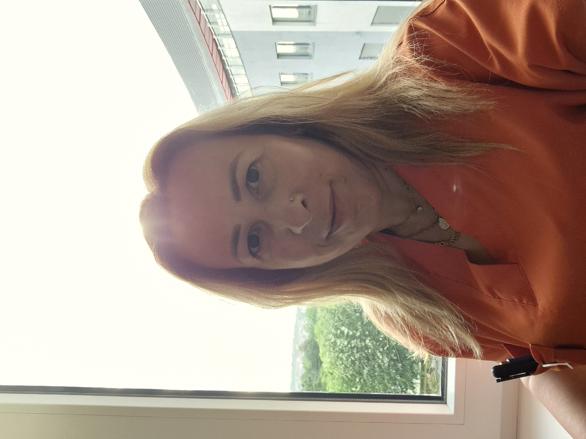 A woman with long, light blonde hair is captured in a selfie or portrait, looking directly at the camera with a gentle, slight smile. She has a small silver stud piercing on her left nostril. She is wearing an orange or burnt orange top, possibly a uniform or scrubs, which includes a collar and a breast pocket. A dark object, resembling a pen or a small device, is clipped to this pocket. Around her neck, she wears a thin silver-colored necklace with a small heart-shaped pendant.
The setting is indoors, next to a large window. Through the window, a bright, possibly overcast, daytime sky dominates the upper part of the background. Below the sky, a modern building with several rectangular windows is visible. Further down, lush green foliage, likely trees or a park, extends towards the horizon, suggesting a view over a cityscape or natural landscape. A white window frame and sill are prominent in the foreground, framing the outdoor scene. The image conveys a serene atmosphere, taken during daylight hours. No text is visible.