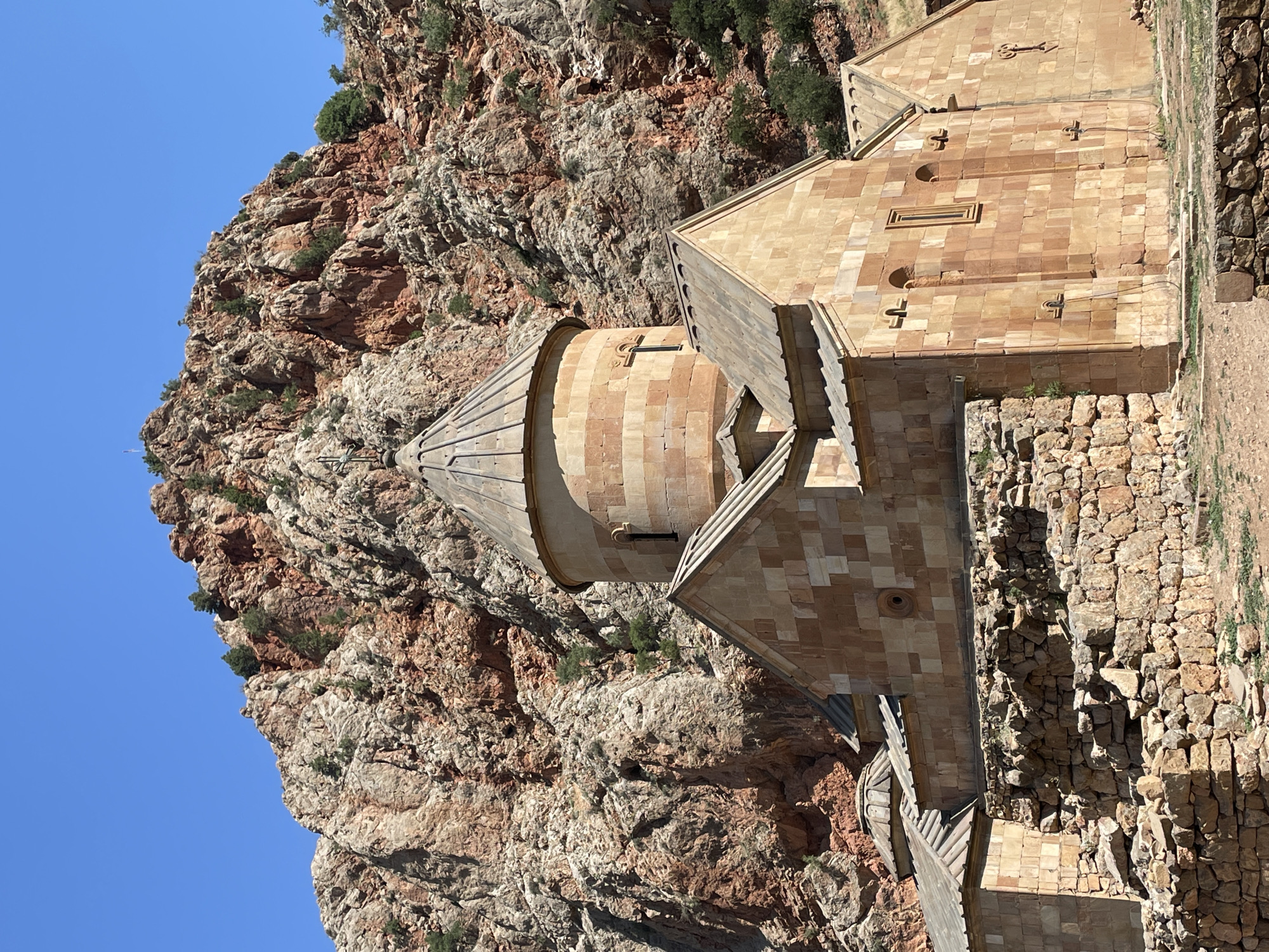 The image displays the Noravank Monastery, an Armenian Apostolic church located in the Republic of Armenia. The monastery is situated in a rocky, mountainous landscape with a clear blue sky. The main structure is built of reddish-brown stone, featuring multiple domed roofs, arched windows, and decorative carvings. In the foreground, a partially ruined stone wall surrounds the monastery complex. Sparse vegetation, including small trees, grows on the rocky slopes behind the church. The scene appears to be captured during daylight, with strong sunlight casting shadows on the buildings and rocks. There are no people visible in the image.