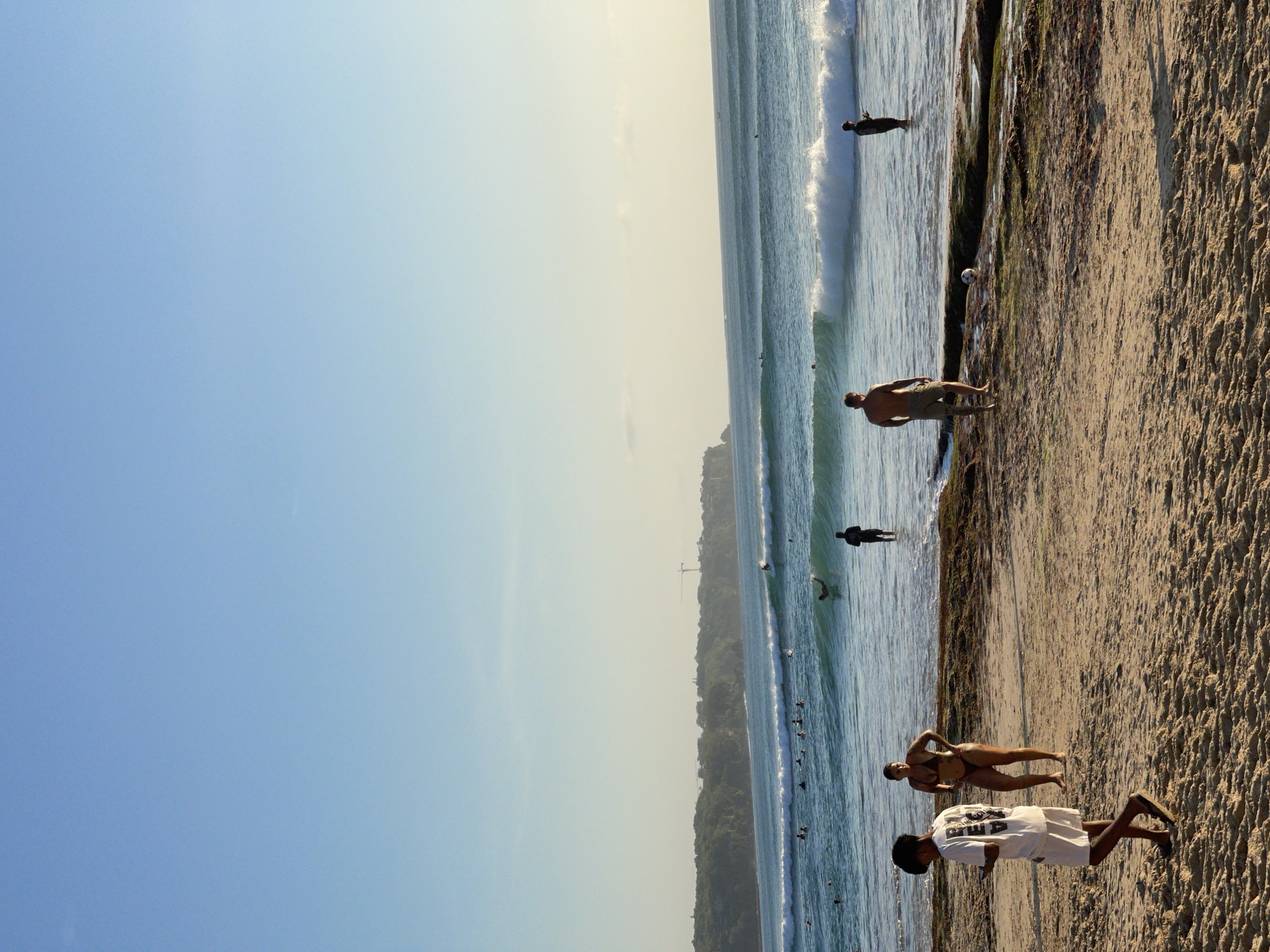 The image depicts a beach scene in Kuta Selatan, Indonesia, with a clear blue sky dominating the upper portion. In the foreground, a sandy beach is visible, with a few people walking and standing. A young person in a white t-shirt and shorts walks from left to right, looking at their phone. To their right, a person in a dark bikini stands facing away from the camera. Further back, a shirtless man in shorts stands on a rocky outcrop in the shallow water, facing the ocean.
The ocean is a prominent feature, with several lines of waves breaking as they approach the shore. Surfers can be seen in the distance, some on their boards and others in the water. The water appears clear and blue-green.
In the background, a lush green cliff face forms the coastline, dotted with some structures. A construction crane is visible atop the cliff, indicating ongoing development. The overall impression is of a popular beach area during daylight hours, likely late afternoon given the quality of light.