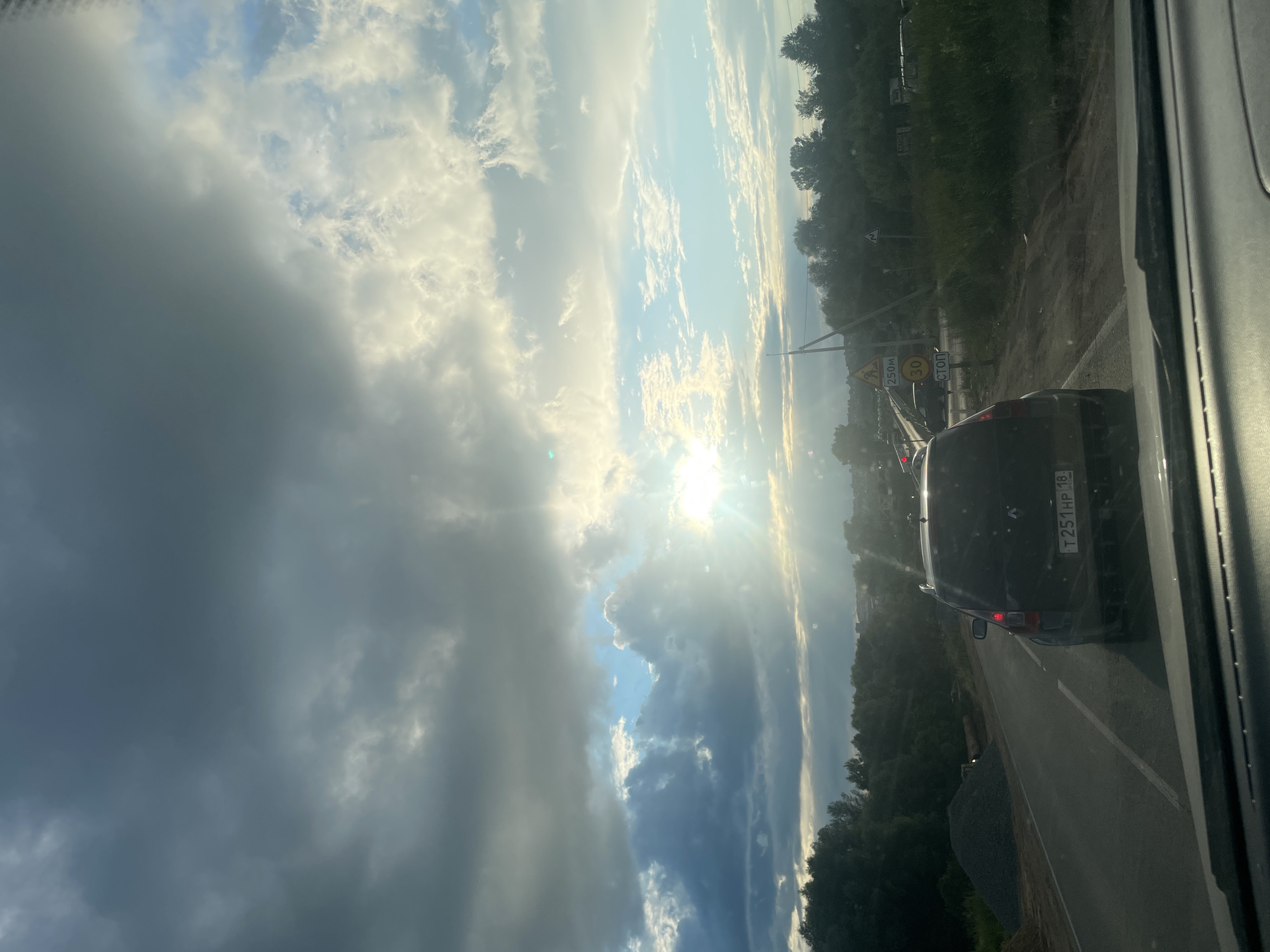 The image depicts a view from inside a vehicle, looking out at a road and a dramatic sky. The scene is set in Гавриловка, Russia, and it appears to be late afternoon or early evening due to the low angle of the sun. The sky is filled with a mixture of dark, heavy clouds and bright, sunlit patches, suggesting a dynamic weather pattern, possibly the end of a storm or approaching twilight. The sun's rays are breaking through the clouds, creating a lens flare and illuminating the scene.
On the right side of the frame, a dark-colored SUV is visible in traffic on a paved road. The vehicle has a license plate that reads "T251HP 18". There are road signs visible, including a yellow warning sign with a triangle and a zigzag line, and a circular sign with "30" inside, indicating a speed limit. Another sign reads "СТОП," which translates to "STOP." The road is bordered by lush green trees on the right and a somewhat unkempt embankment on the left, with piles of gravel.
The overall mood is somewhat somber due to the heavy clouds, but also hopeful and bright due to the sunlight breaking through. The image captures a moment of travel on a rural or suburban road, under a striking sky, in what appears to be a transitional time of day.