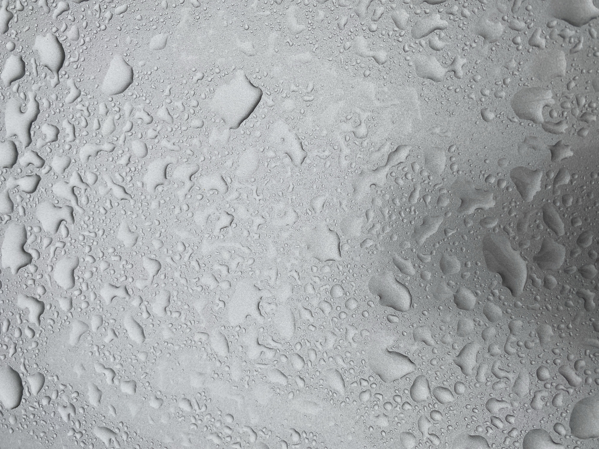 The image depicts a surface covered in water droplets. The droplets vary in size, ranging from tiny beads to larger, irregular shapes. The surface appears to be a metallic material, possibly with a matte finish, considering its uniform gray tone. There are no people, actions, or interactions visible. There's no information related to the location in the city of Noviy, Russia.