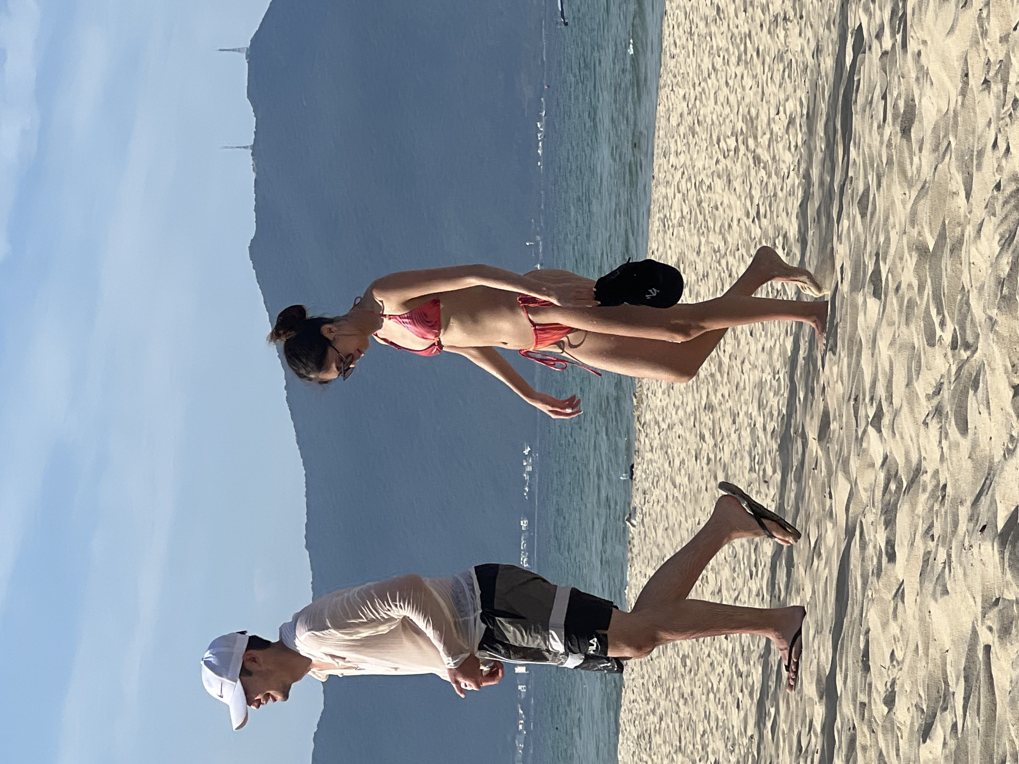 Two people are walking on a sandy beach in Đà Nẵng, Vietnam. The person on the left is a man wearing a white baseball cap, a translucent white long-sleeved shirt, and dark shorts with a white stripe and "FILA" logo. He is walking to the left. The person on the right is a woman wearing a red and pink patterned bikini and sunglasses. She is walking to the left and carrying a black hat in her right hand. The sea is visible behind them, with a hilly landscape in the distance. The sky is clear with some clouds. The sand is light-colored and textured with shadows.