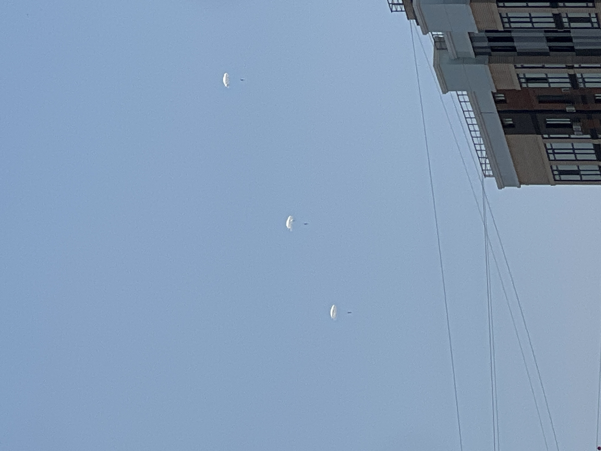 A clear blue sky is visible above a modern apartment building. Three white parachutes are suspended in the sky, each with a dark figure beneath it, indicating parachutists. The building, located in the city of Krasnodar, Russia, has a contemporary design with a mix of beige, brown, and grey facades, and numerous windows. Power lines are strung across the scene, obscuring parts of the sky and the building.