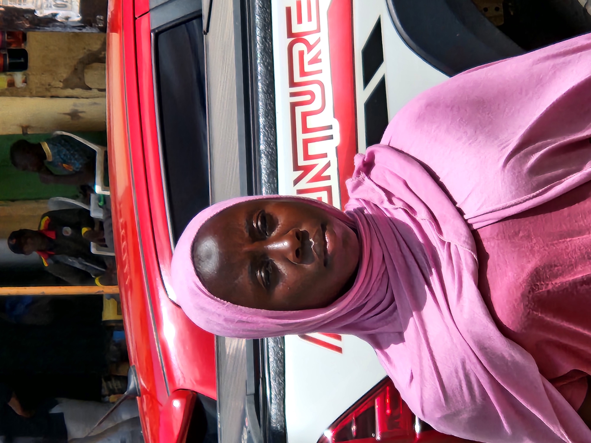 A medium shot captures a Black woman wearing a pink hijab and a pink top. She is standing in front of a red vehicle with a white truck bed that has "NATURE" written in red letters. To her left, the red vehicle appears to be a car. In the background, a man in a dark uniform with yellow trim is seated in front of a building. To the right of the seated man, another Black man wearing a blue and yellow shirt is also seated. The scene is outdoors, suggesting a street or market setting in Jos, Nigeria. The lighting is bright, indicating daytime.