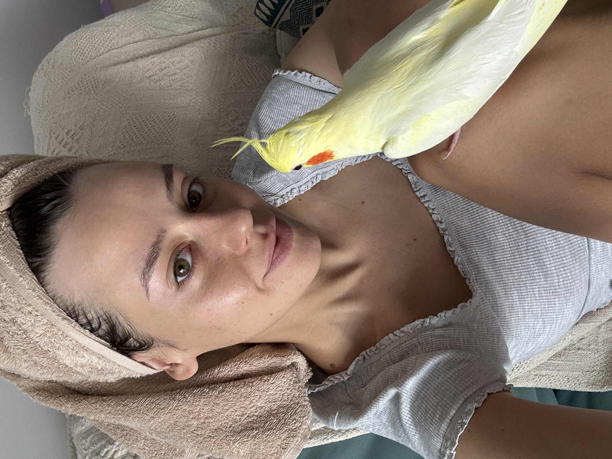 The image shows a close-up of a young woman with a tan towel wrapped around her head, her hair wet. She is wearing a grey t-shirt with ruffled edges. She is holding a yellow cockatiel on her shoulder, which is perched on her arm. The bird has a red patch on its cheek and a crest on its head. The background appears to be an indoor setting with a textured blanket. There are no identifiable landmarks or elements that would indicate a specific location in the city of Krasnoyarsk, Russia.